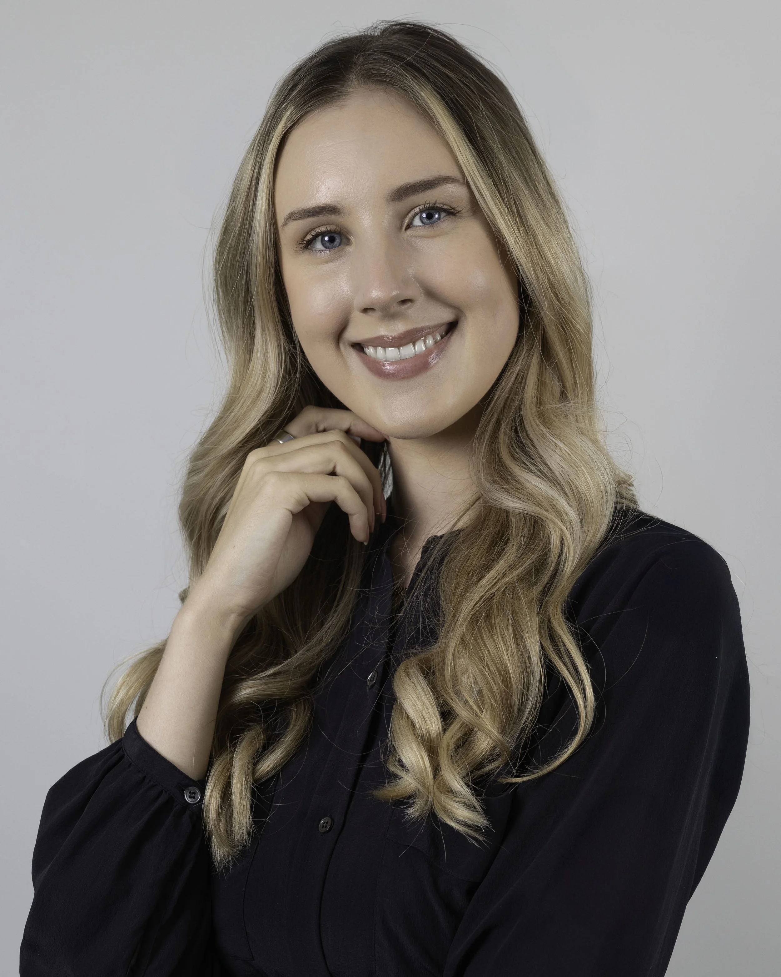 A smiling woman with long, wavy blonde hair, wearing a black shirt, posing with her right hand touching her chin against a plain grey background.