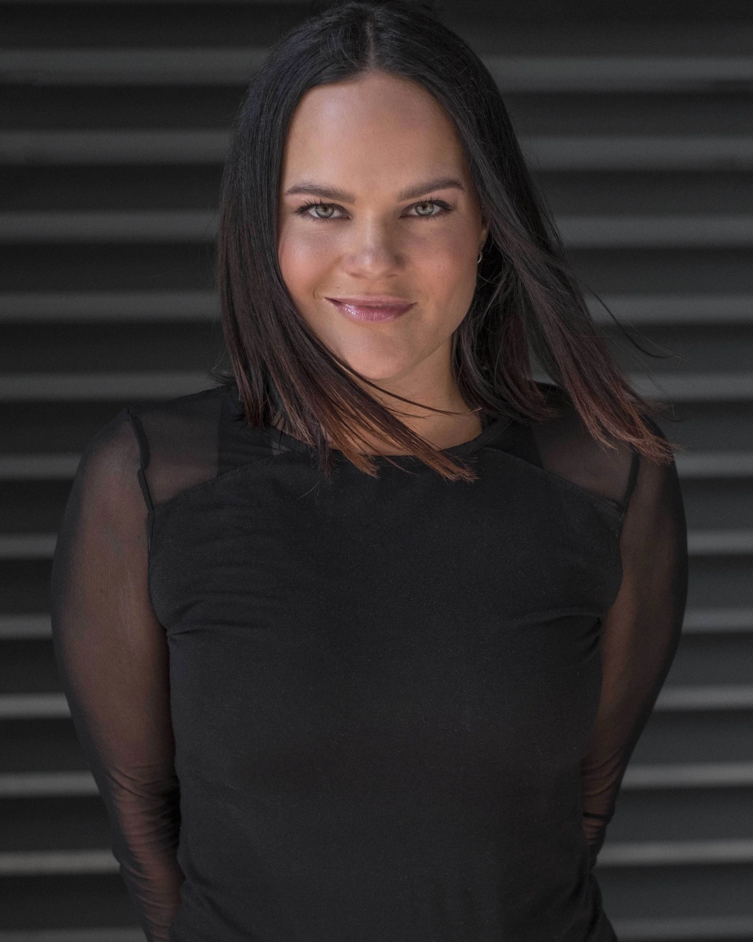 A woman with shoulder-length dark hair and blue eyes, wearing a black top with sheer sleeves, standing in front of a dark, striped background.