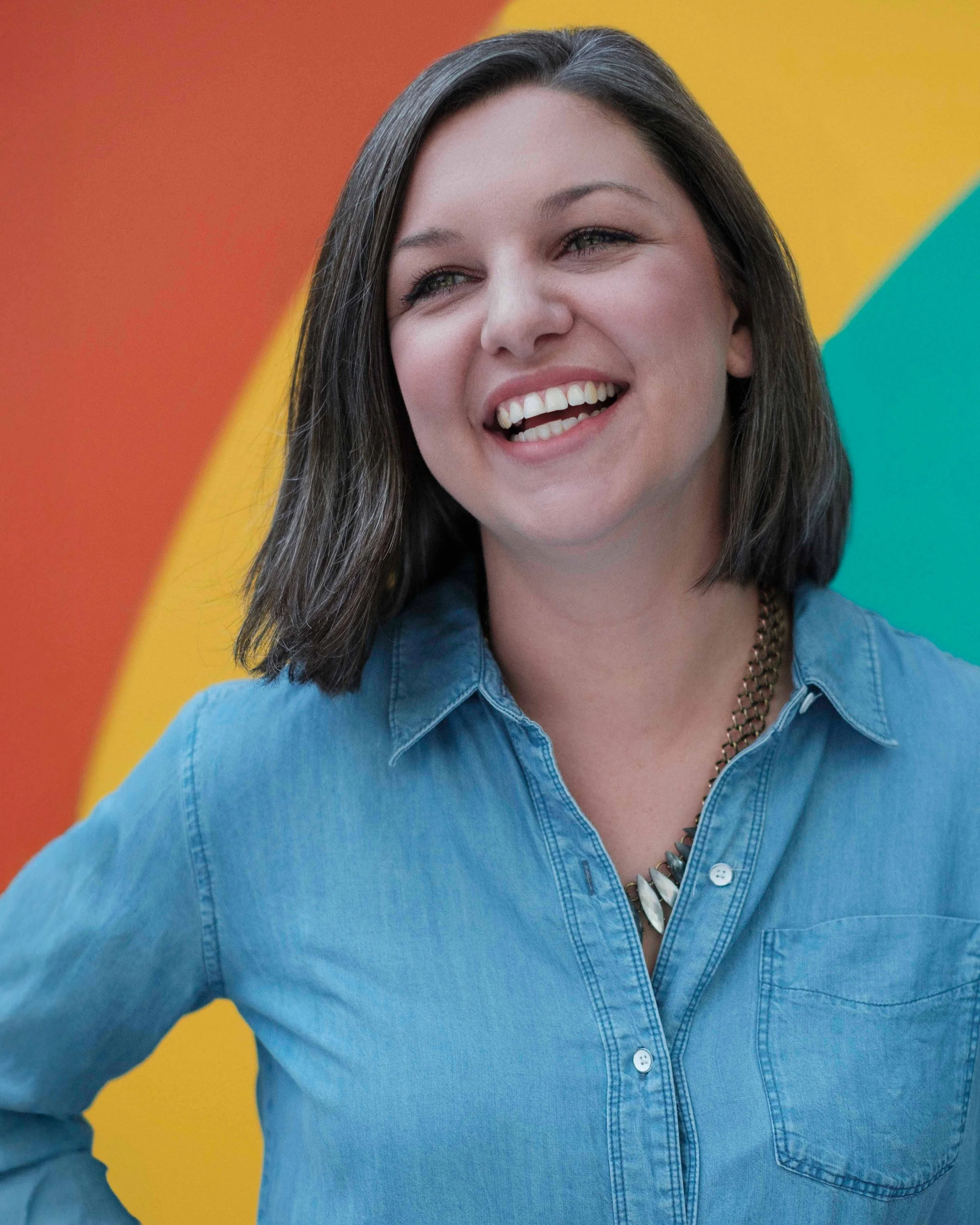 A woman with shoulder-length dark hair smiling and wearing a denim shirt against a colorful background with red, yellow, and teal sections.