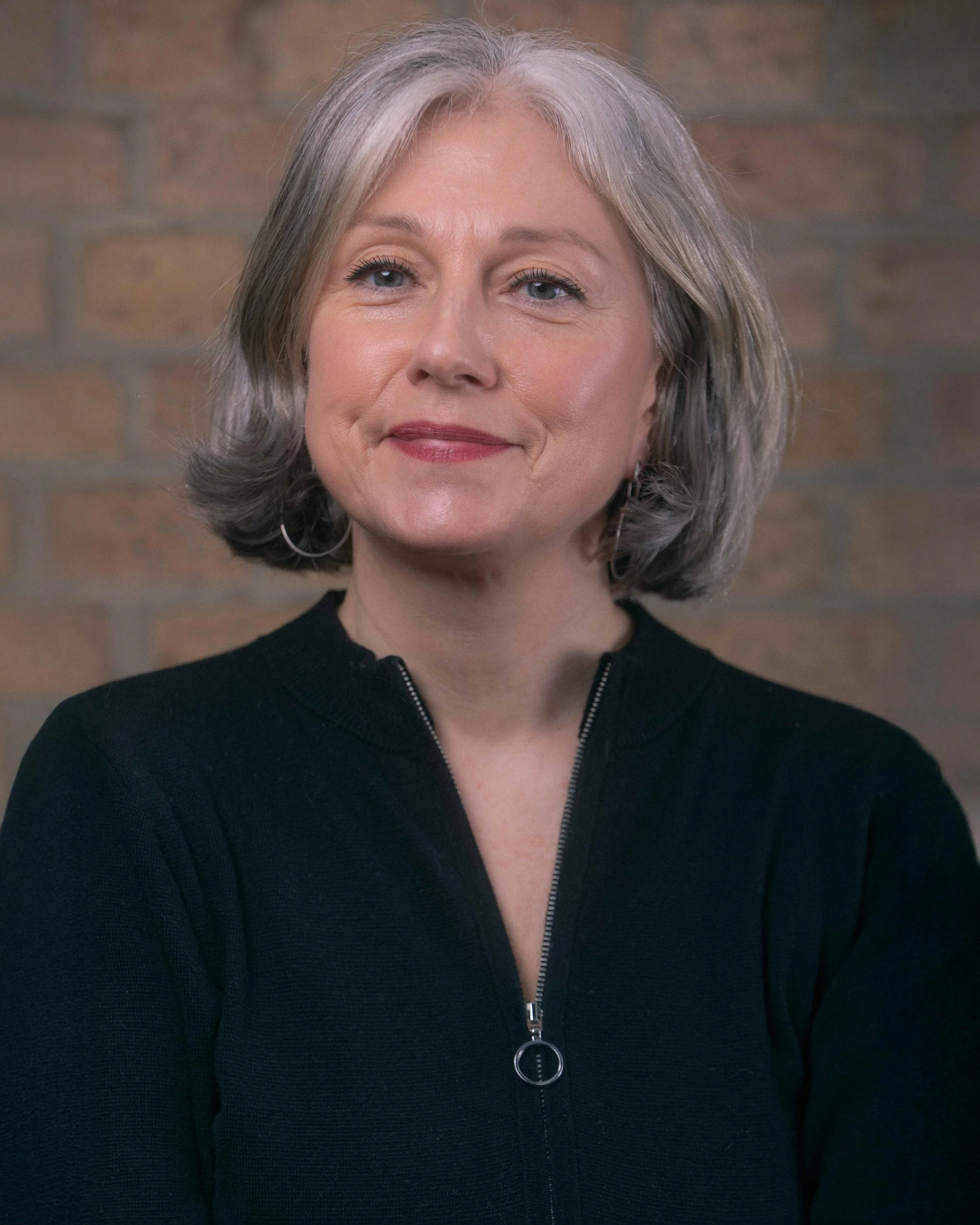 A woman with short gray hair, wearing a black zip-up top, smiling slightly, against a brick wall background.