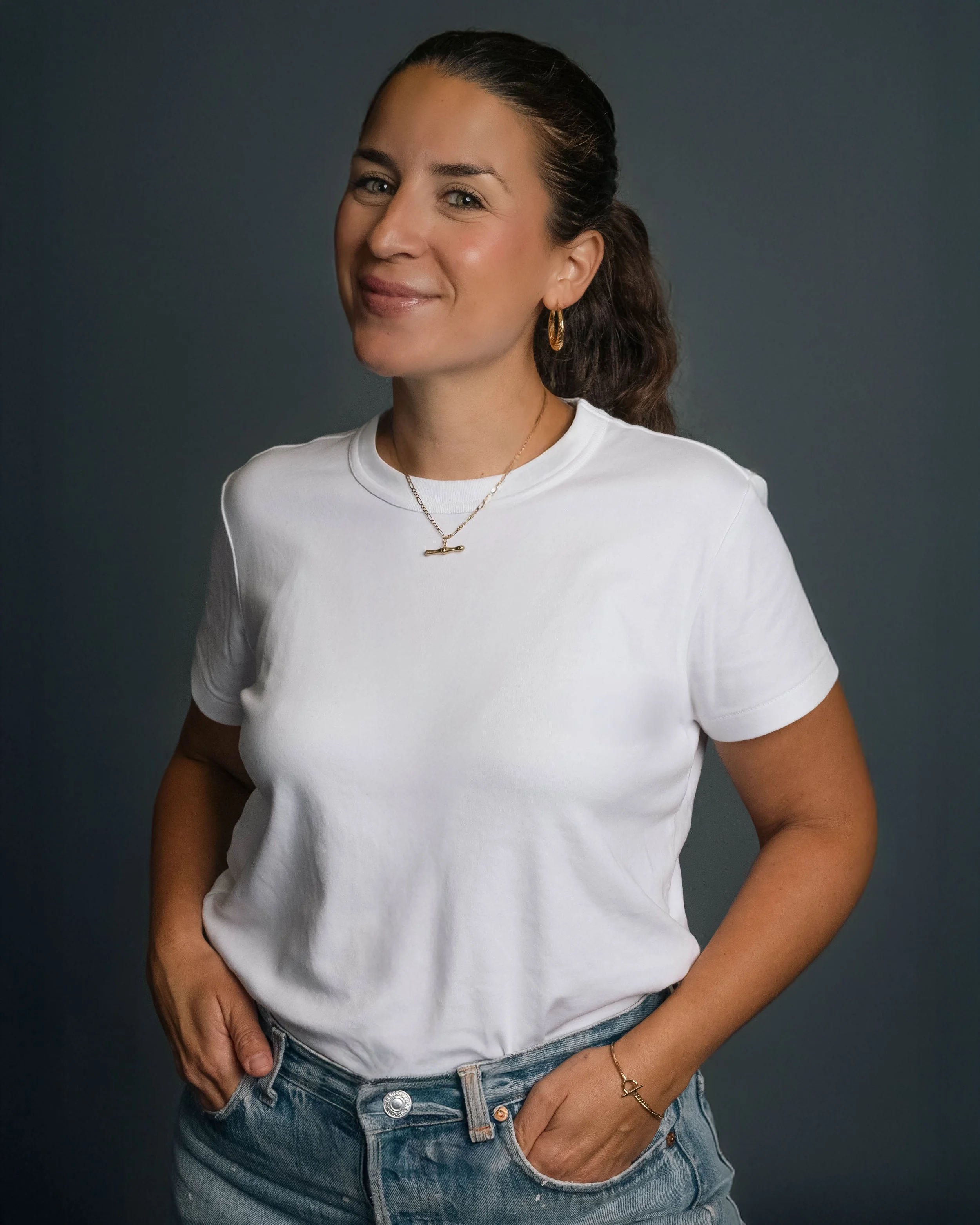 A woman wearing a white t-shirt and jeans, smiling with her hands in her pockets, against a dark background.
