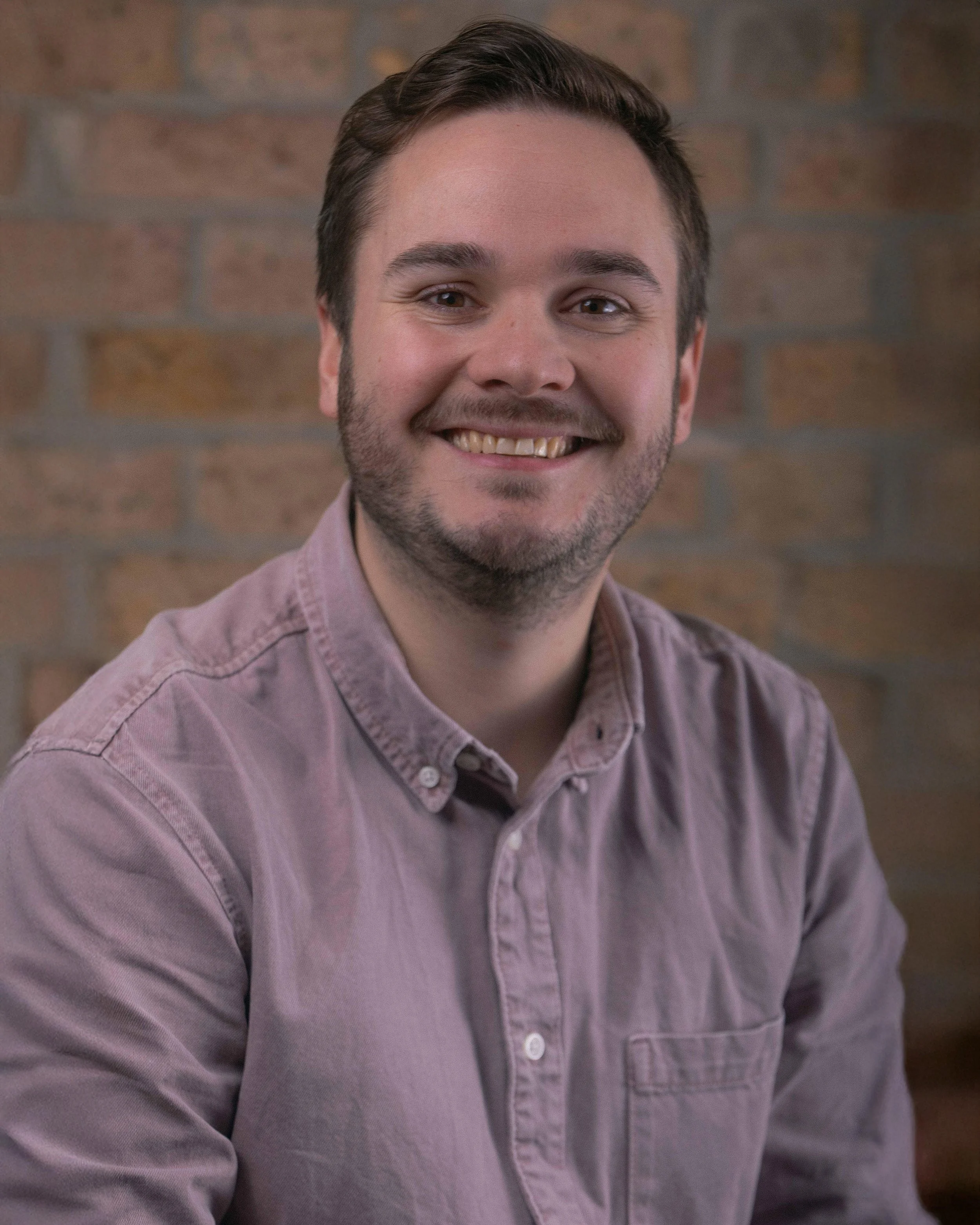 A smiling man with short brown hair and facial hair, wearing a light purple button-up shirt, sitting in front of a brick wall.