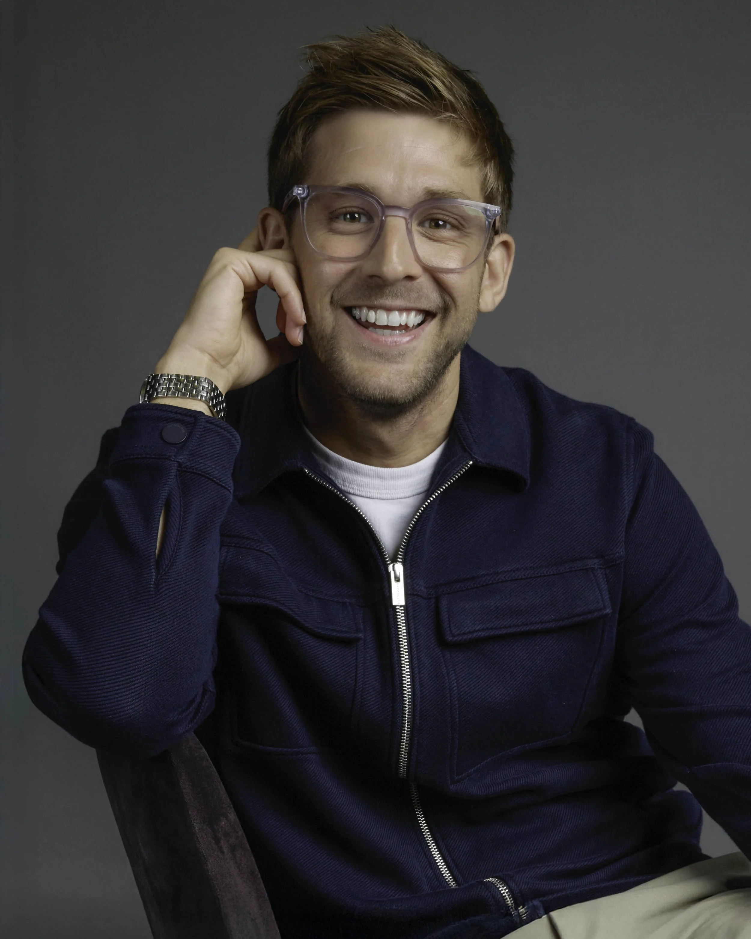 Young man smiling, wearing glasses and navy jacket, sitting on a chair against a grey background.