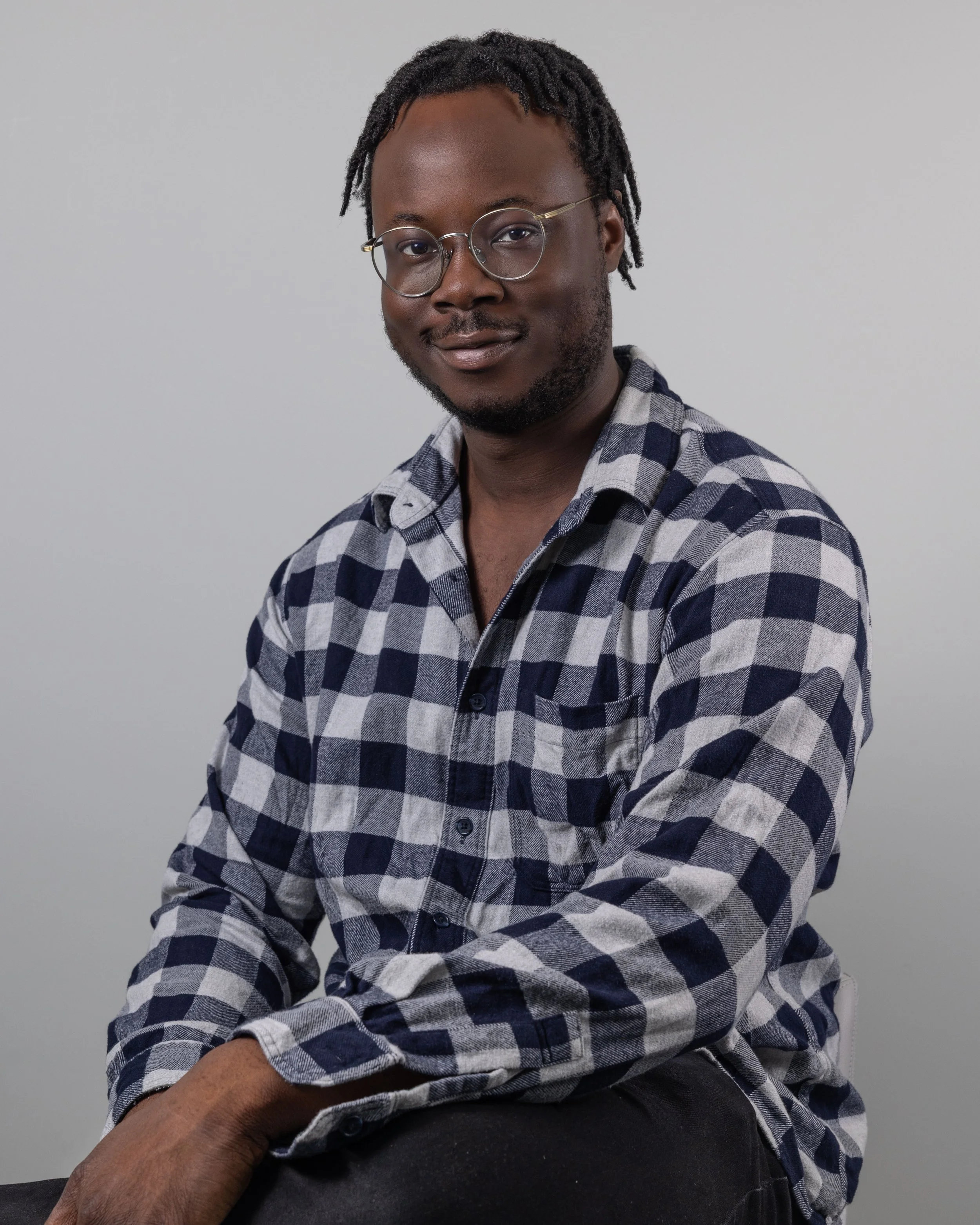 Portrait of a man with glasses, wearing a checkered shirt, sitting against a plain light gray background.