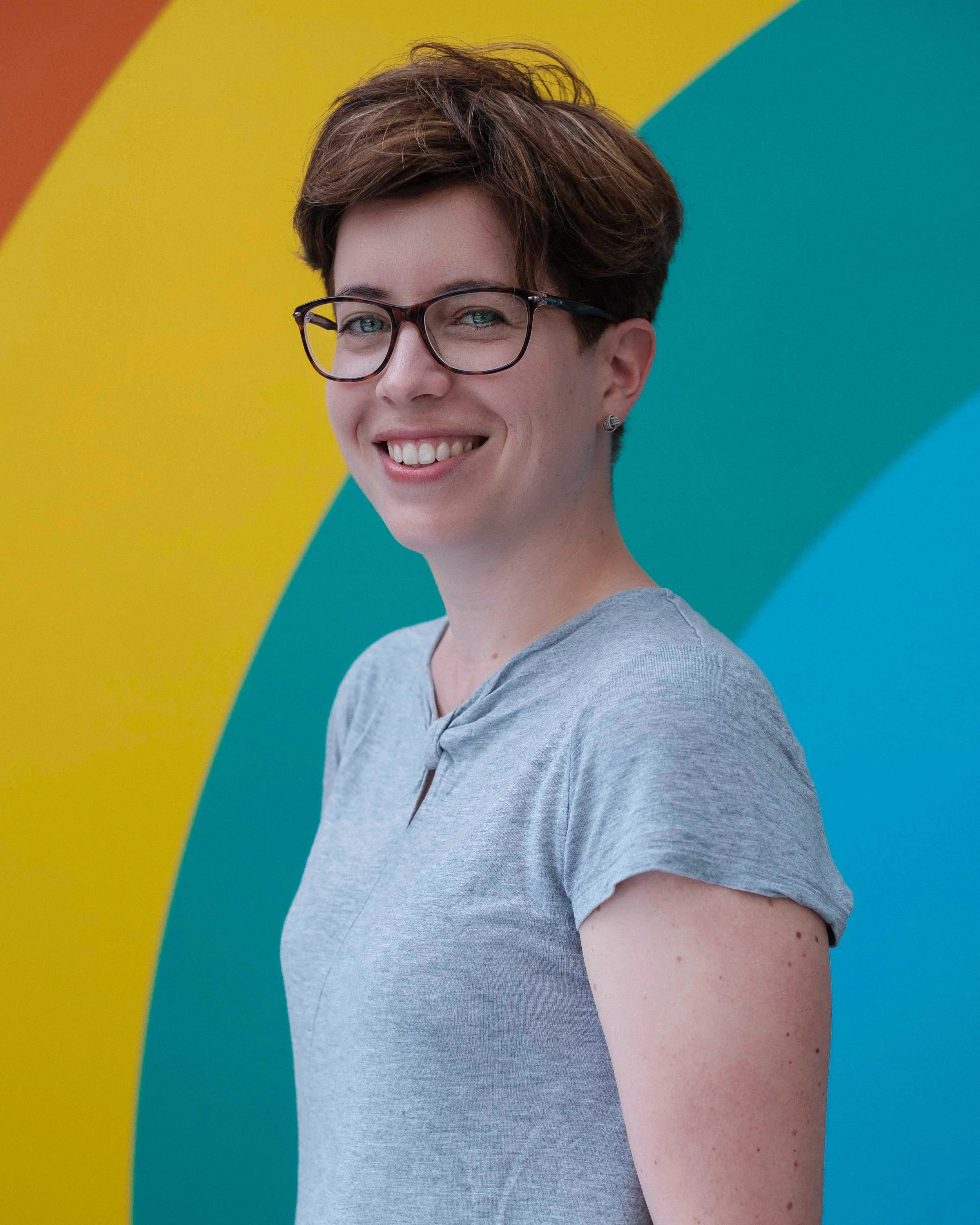 A smiling woman with short brown hair, glasses, and earrings, wearing a light gray T-shirt, standing in front of a colorful, rainbow-striped background.