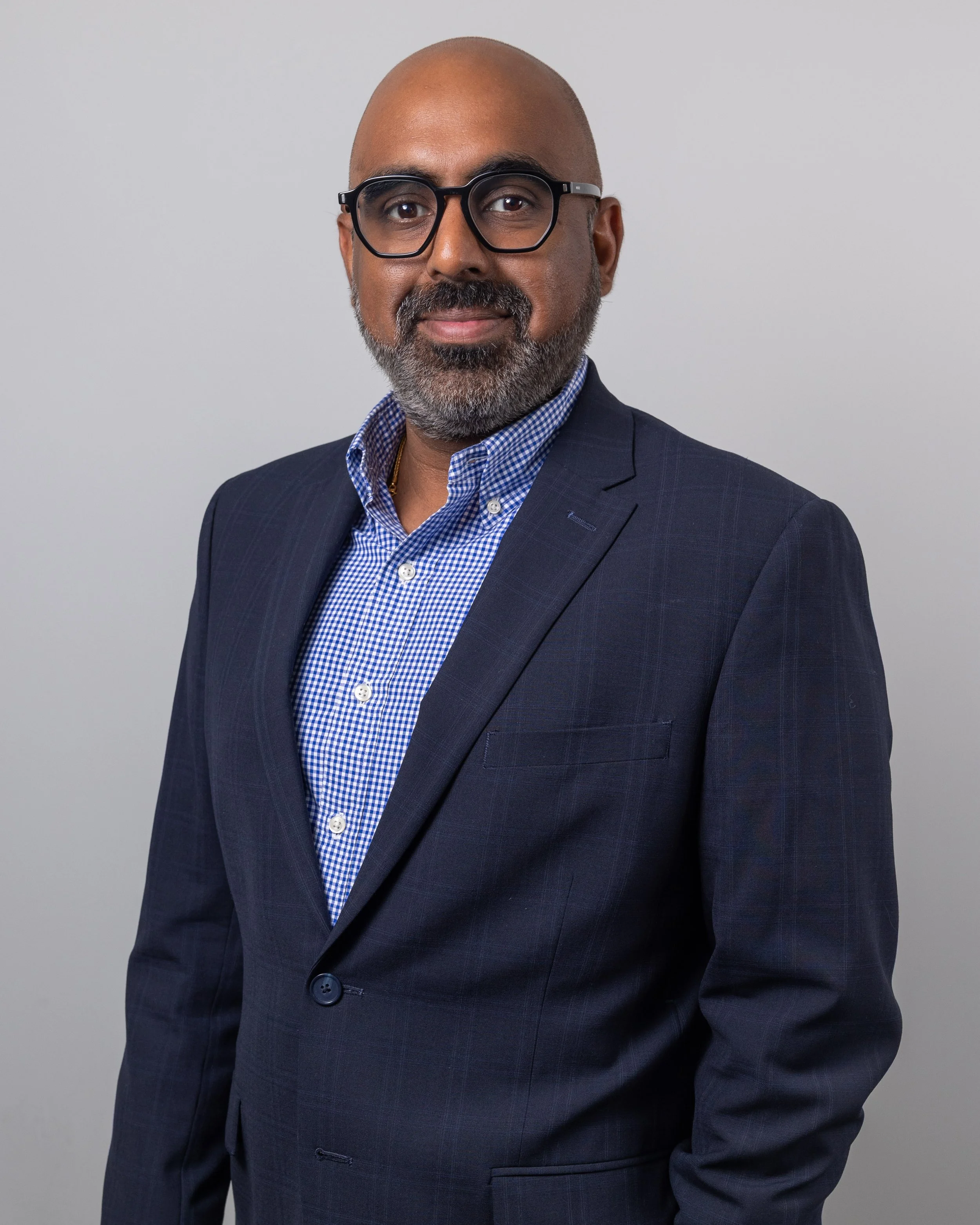 A middle-aged man with a gray beard, bald head, glasses, wearing a dark checkered suit and a blue checkered shirt, smiling slightly against a plain gray background.