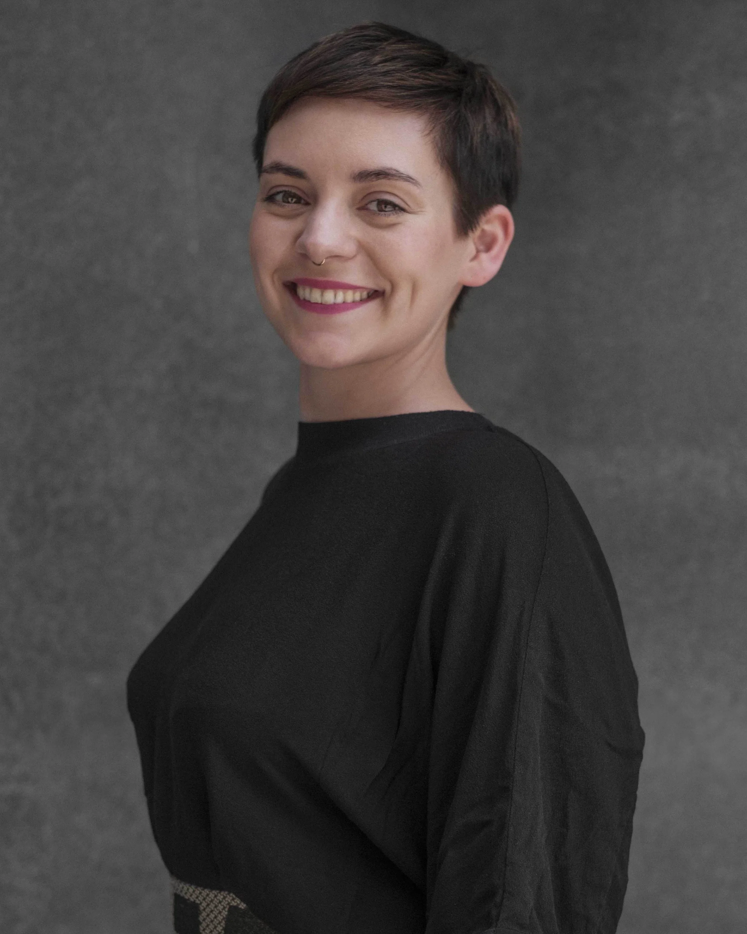 Portrait of a young woman with short dark hair, wearing a black top, smiling against a gray background.