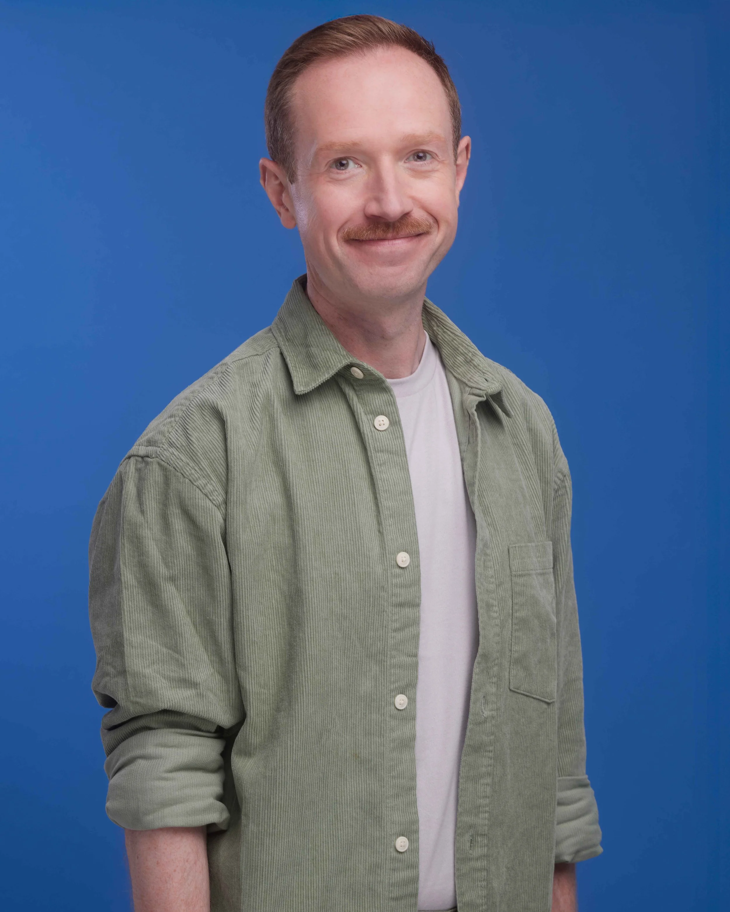 A man with red hair, a mustache, wearing a green button-up shirt over a white t-shirt, standing against a solid blue background, smiling at the camera.