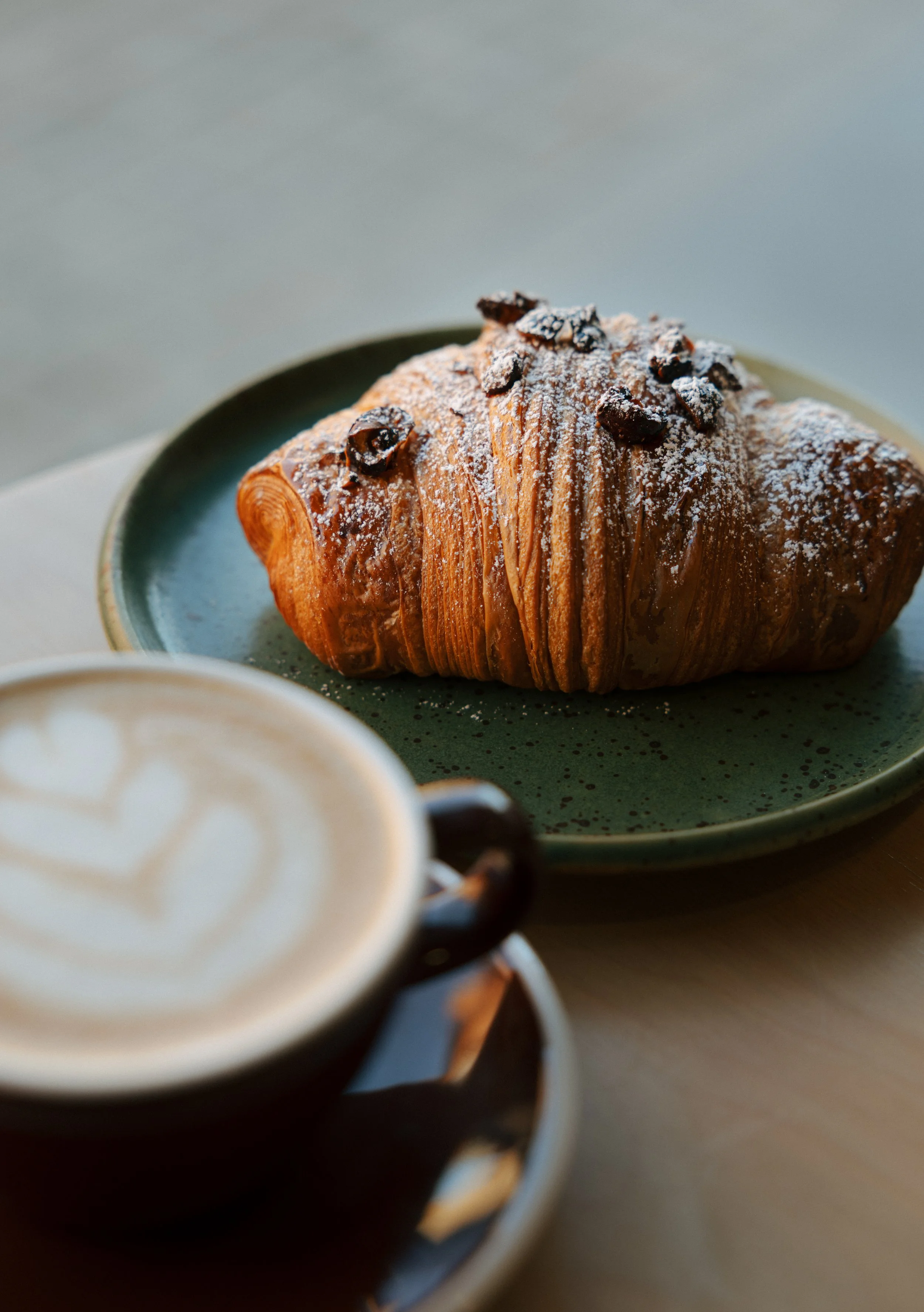 Un croissant en un plato verde acompañado de una taza de café con leche con arte en la espuma.