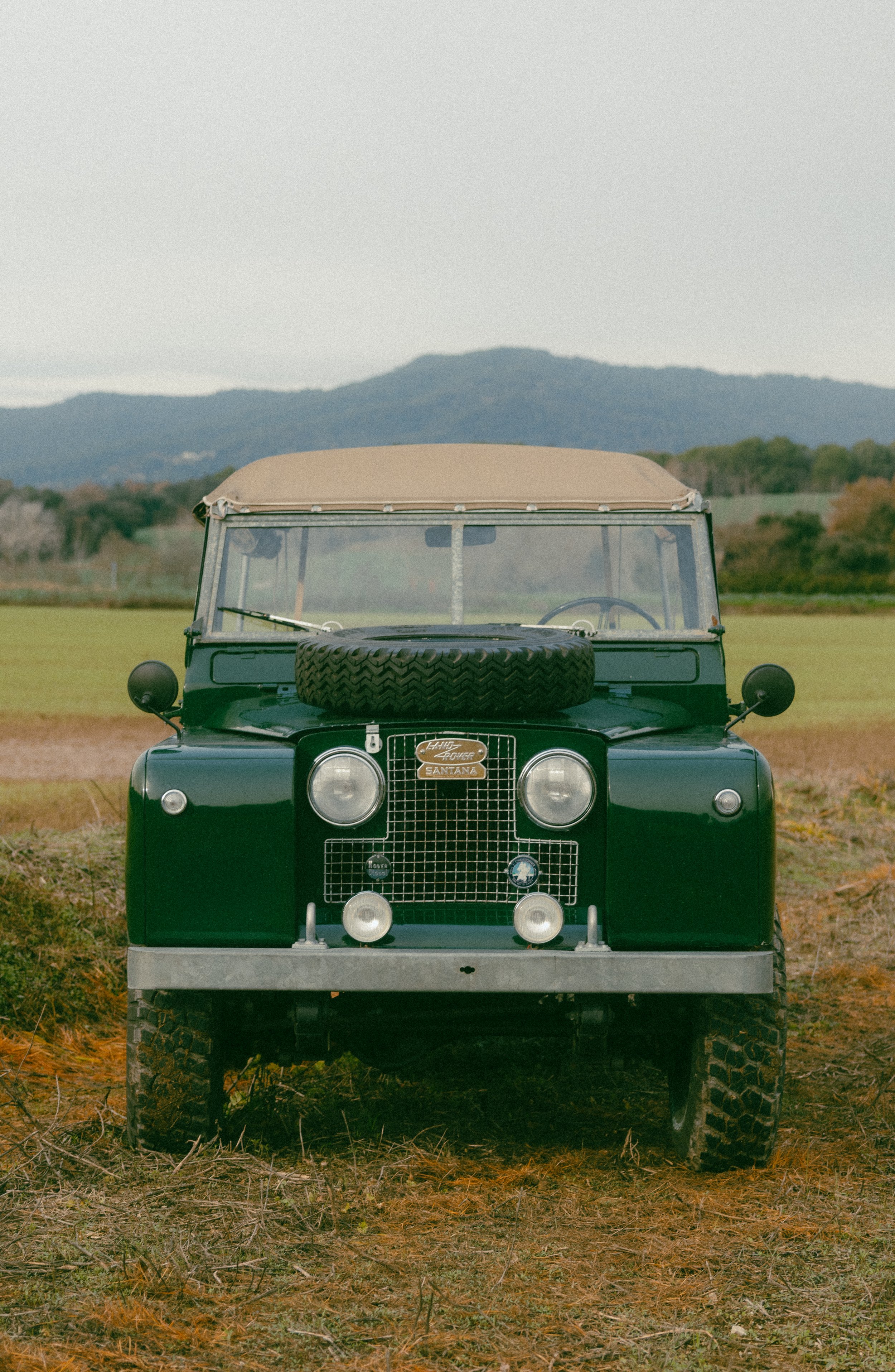 Vehículo clásico de color verde oscuro en un campo con montaña en el fondo.