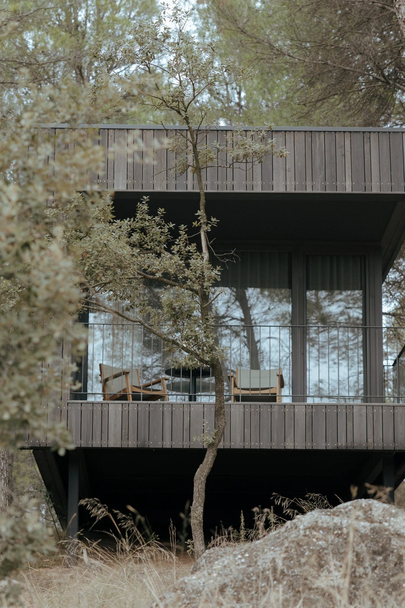 Vista de una casa moderna de dos niveles con balcón de madera y ventanas grandes, rodeada de árboles y naturaleza.