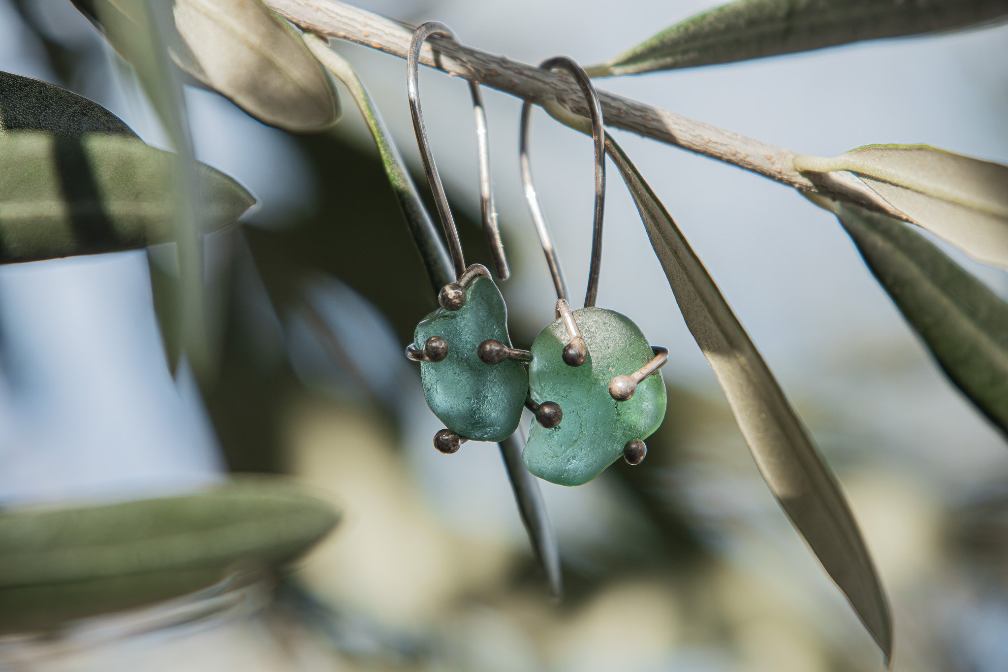 Pendientes de piedra de color verde con detalles de metal, colgados de una rama de árbol con hojas verdes.