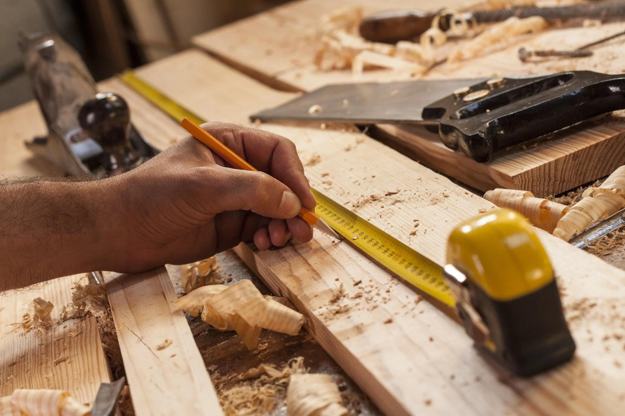 A person measures a wooden plank with a yellow tape measure and a pencil. The workspace is cluttered with wood shavings, chisels, a handsaw, and a table saw.