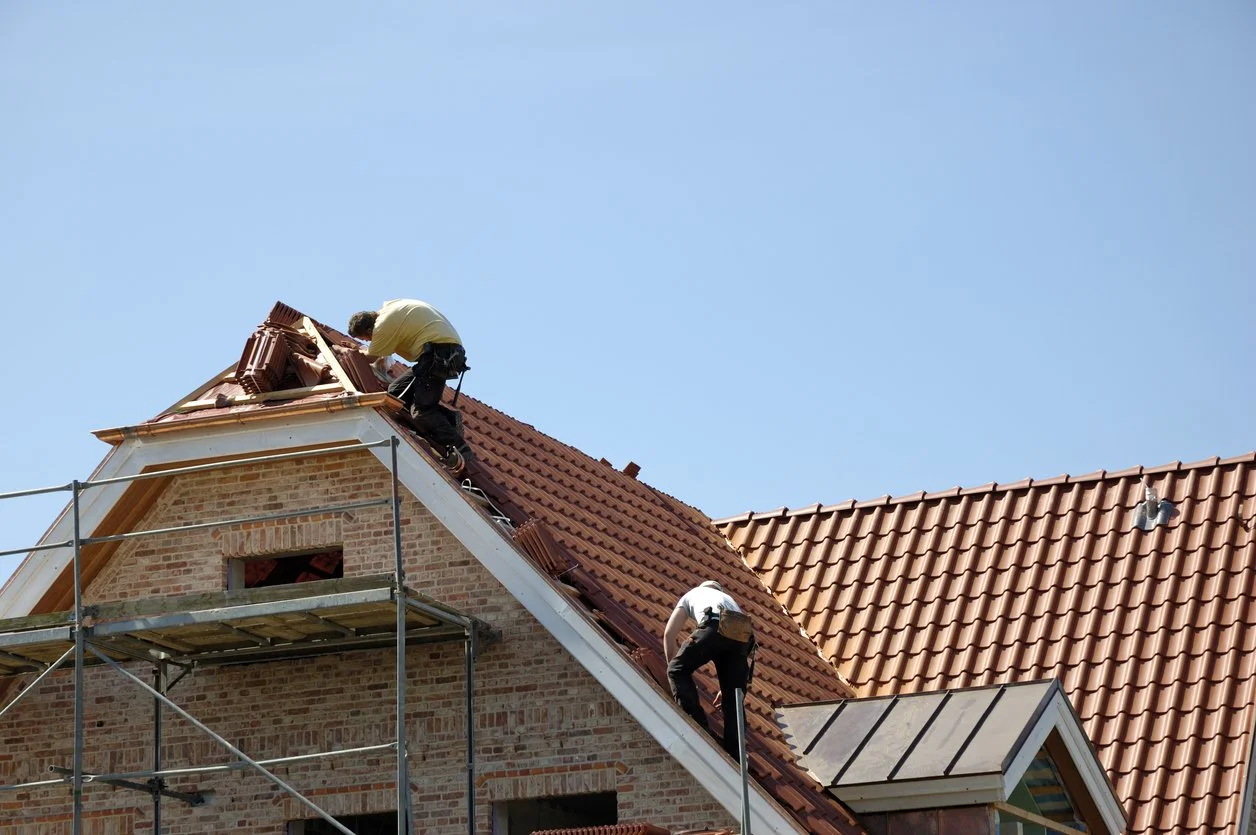 Two workers installing red roof tiles on a brick house with scaffolding outdoors under a clear blue sky.