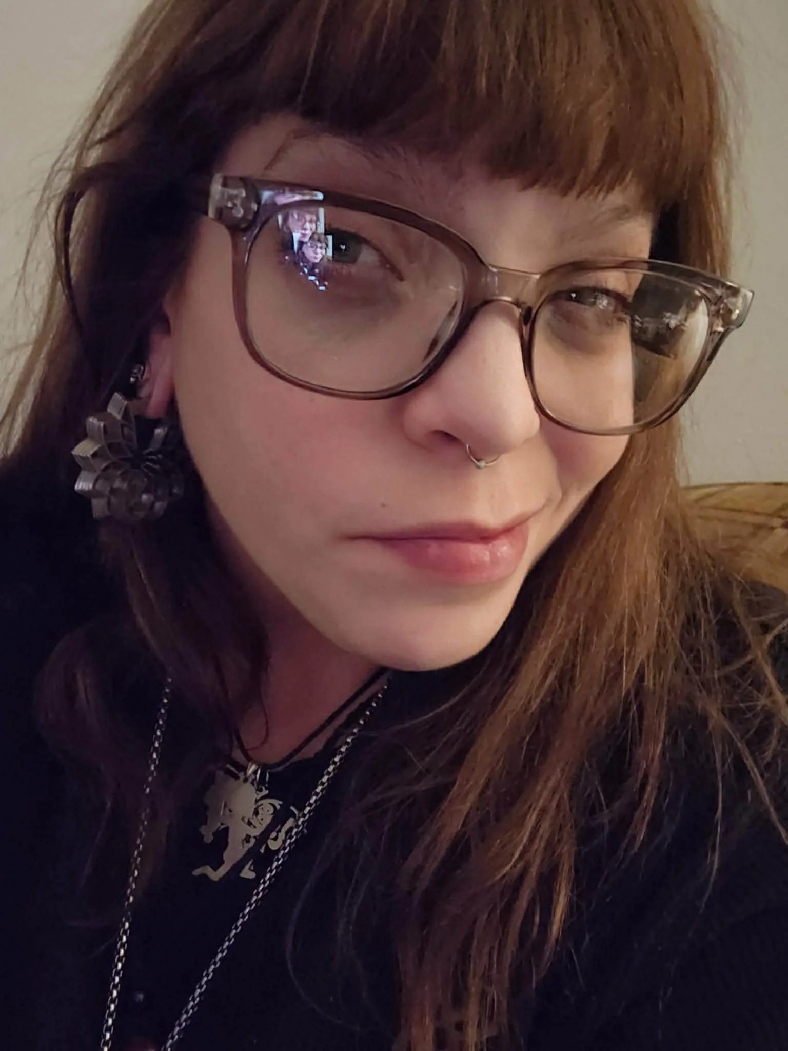 Close-up of a young woman with long, red hair wearing glasses, a septum piercing, a dangling earring, and a black outfit with layered necklaces, smiling softly at the camera.