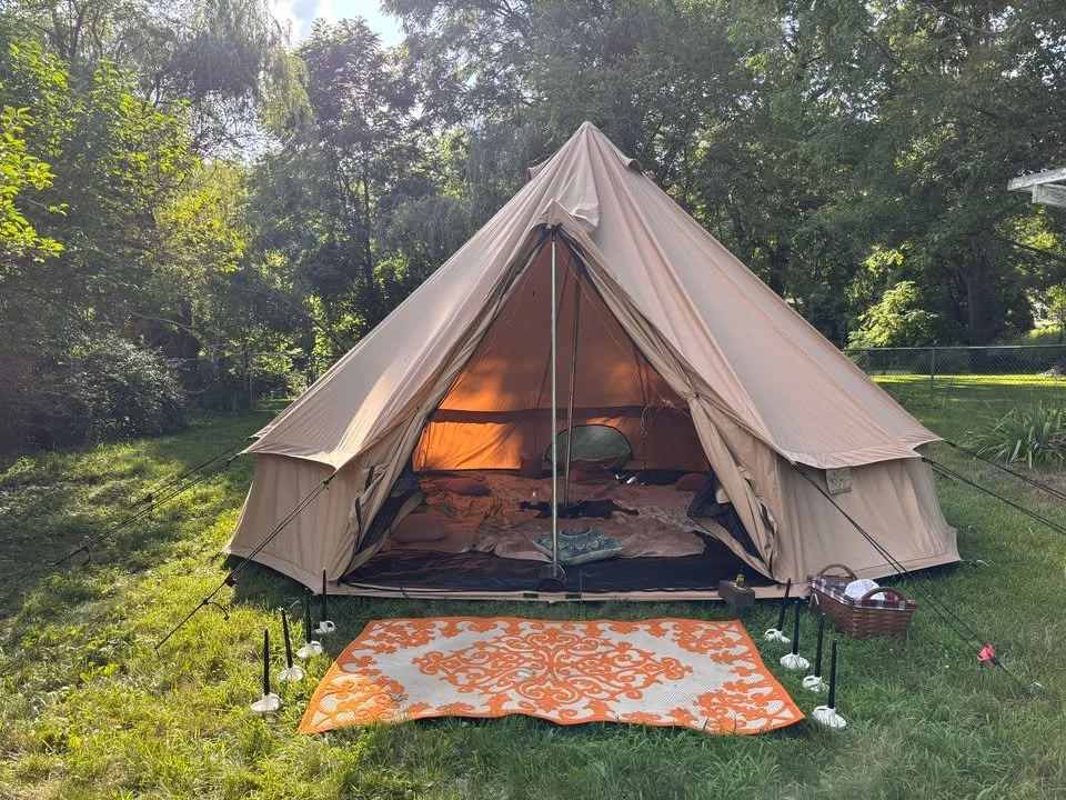A beige camping tent set up on a grassy area surrounded by trees, with an orange and white patterned rug in front and small lights on stakes around it. There is a wicker basket on the right side of the tent.
