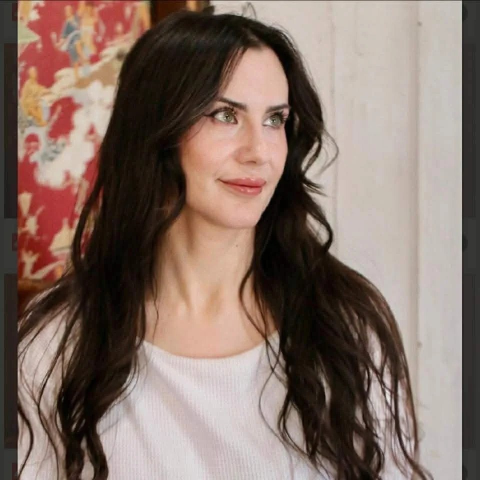 A woman with long, dark wavy hair and light skin, looking to her left and smiling slightly, wearing a white top.