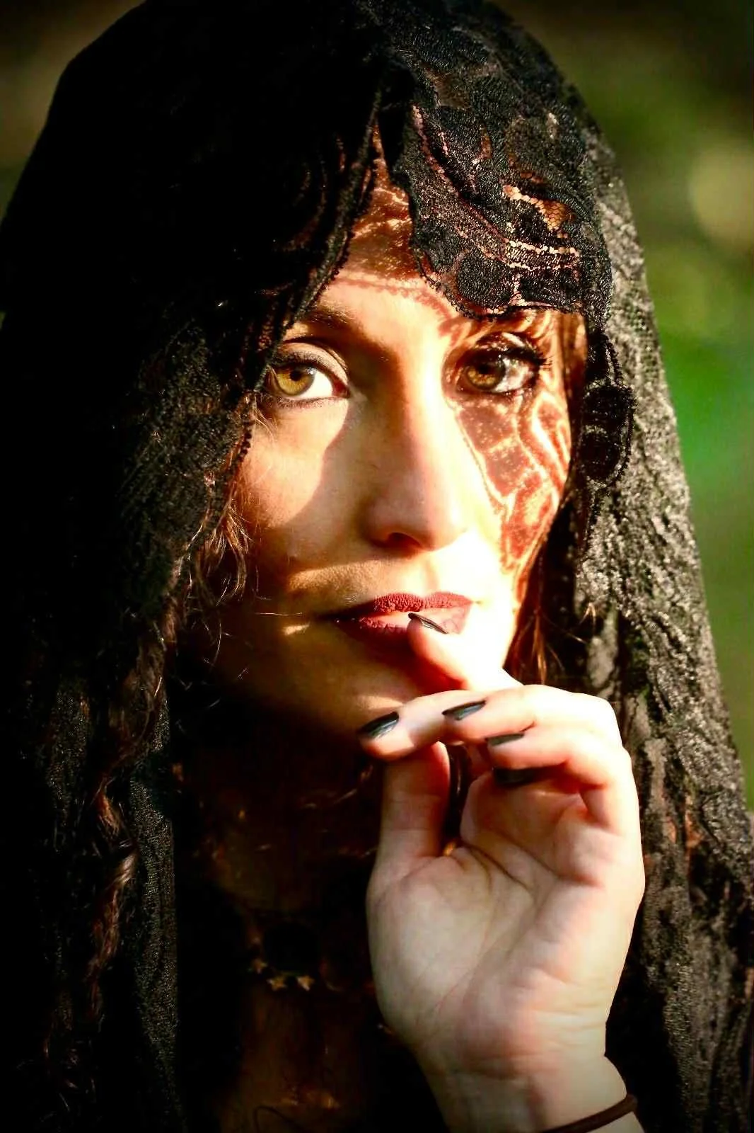 A woman with light brown eyes and dark hair partially covered by black lace fabric, looking thoughtfully with her hand near her mouth.