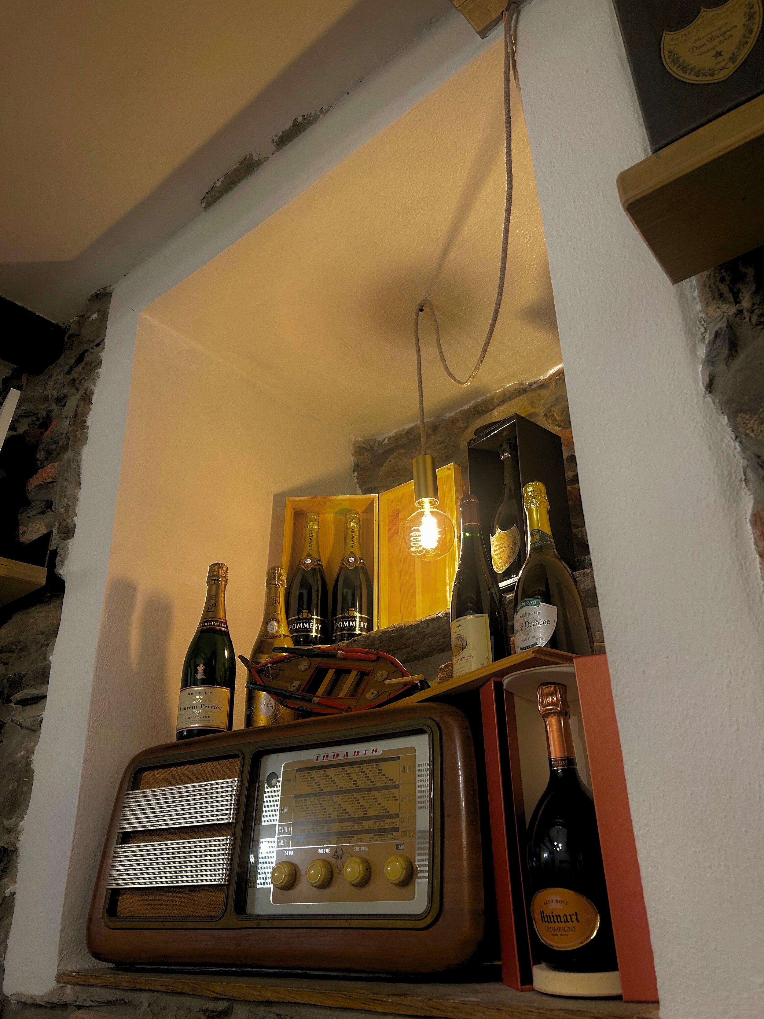 Interno di nicchia con vecchia radio e bottiglie di champagne illuminate da lampadina appesa.