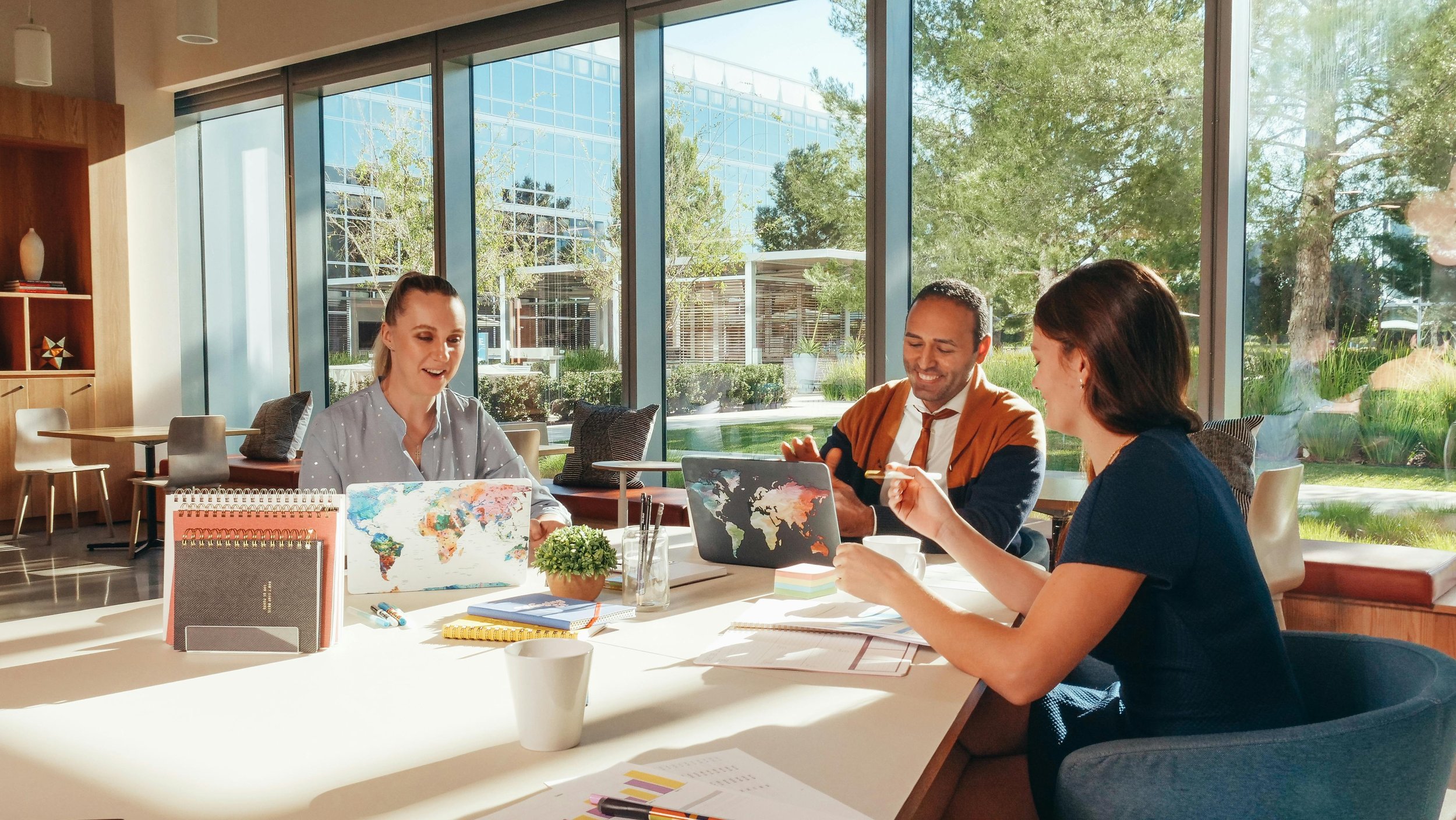 Three professionals in a modern office setting, holding laptops and notebooks, in a bright space with floor-to-ceiling windows overlooking lush greenery, ideal for business hubs, investment networking, and corporate workspaces