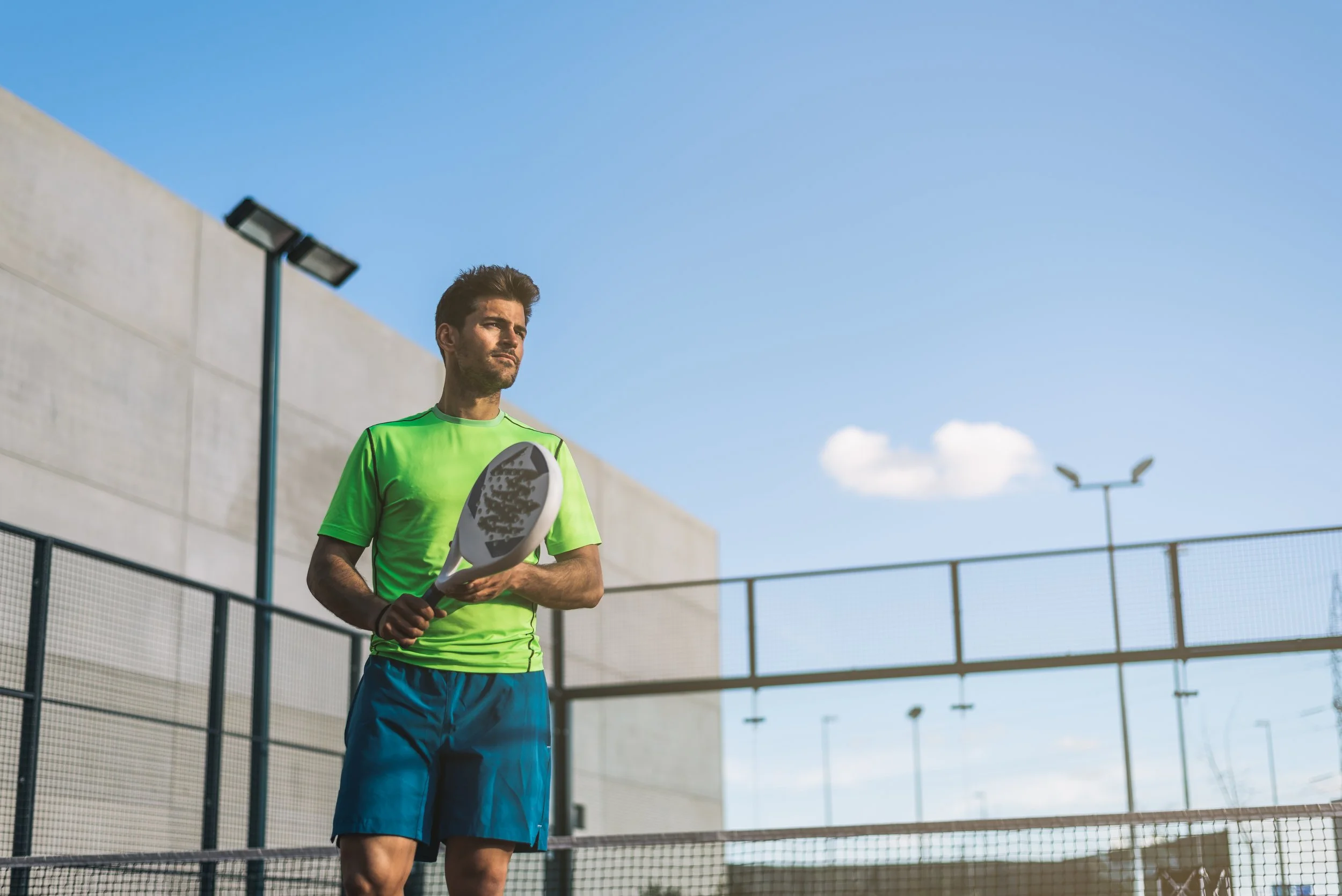 Man playing padel tennis on an outdoor court, wearing a green shirt and blue shorts, highlighting sports facilities in premium real estate investments and resort-style living.