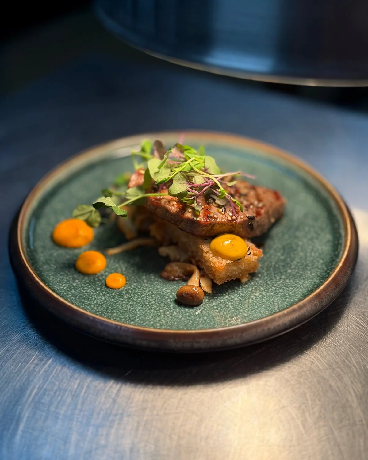 A gourmet dish featuring a piece of meat topped with microgreens, served with small dollops of yellow and orange sauces on a textured green ceramic plate.