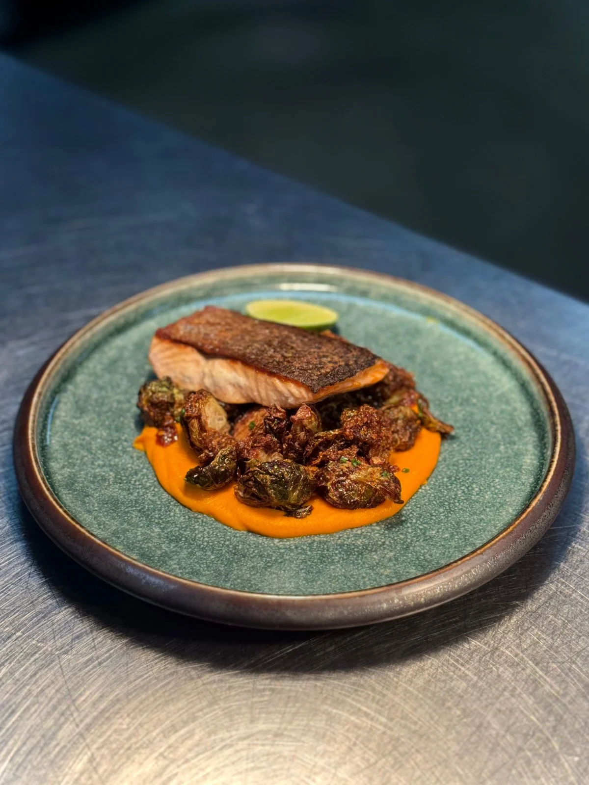 A plated dish featuring a piece of seared fish with crispy skin, served over a bed of roasted Brussels sprouts and topped with bright orange puree and a lime wedge.