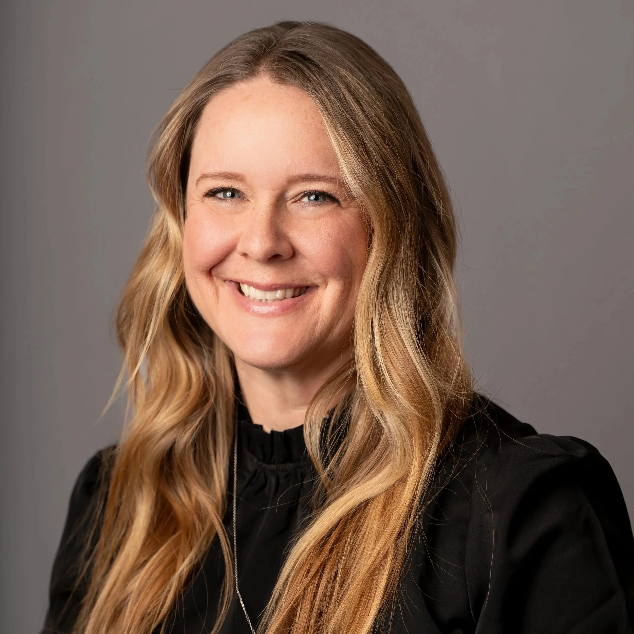 A smiling woman with long, wavy blonde hair wearing a black top against a gray background.