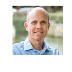 A man with short gray hair and a blue collared shirt smiling outdoors, with a blurred background of water and greenery.