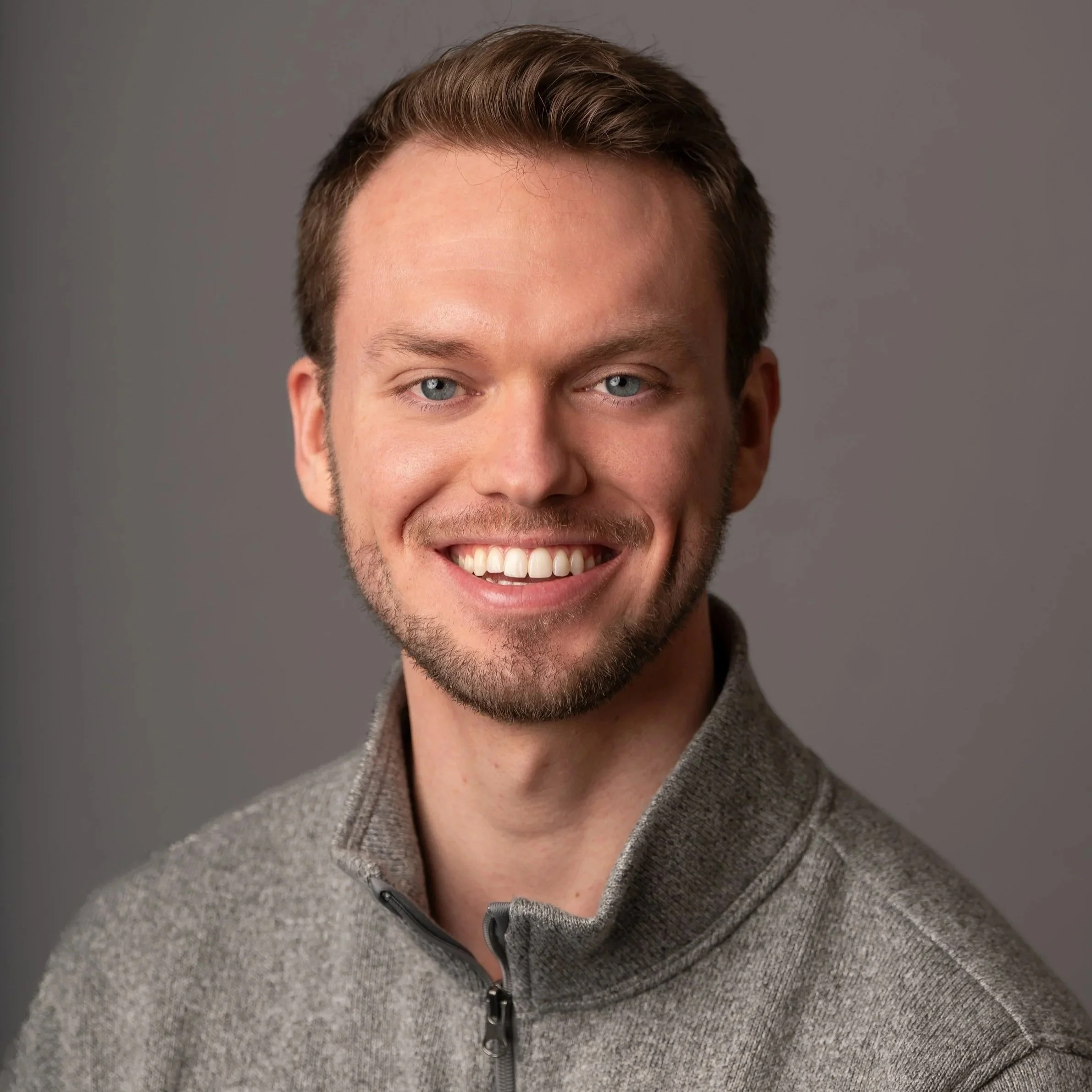 A smiling man with short brown hair and a beard, wearing a gray zip-up sweater, against a gray background.