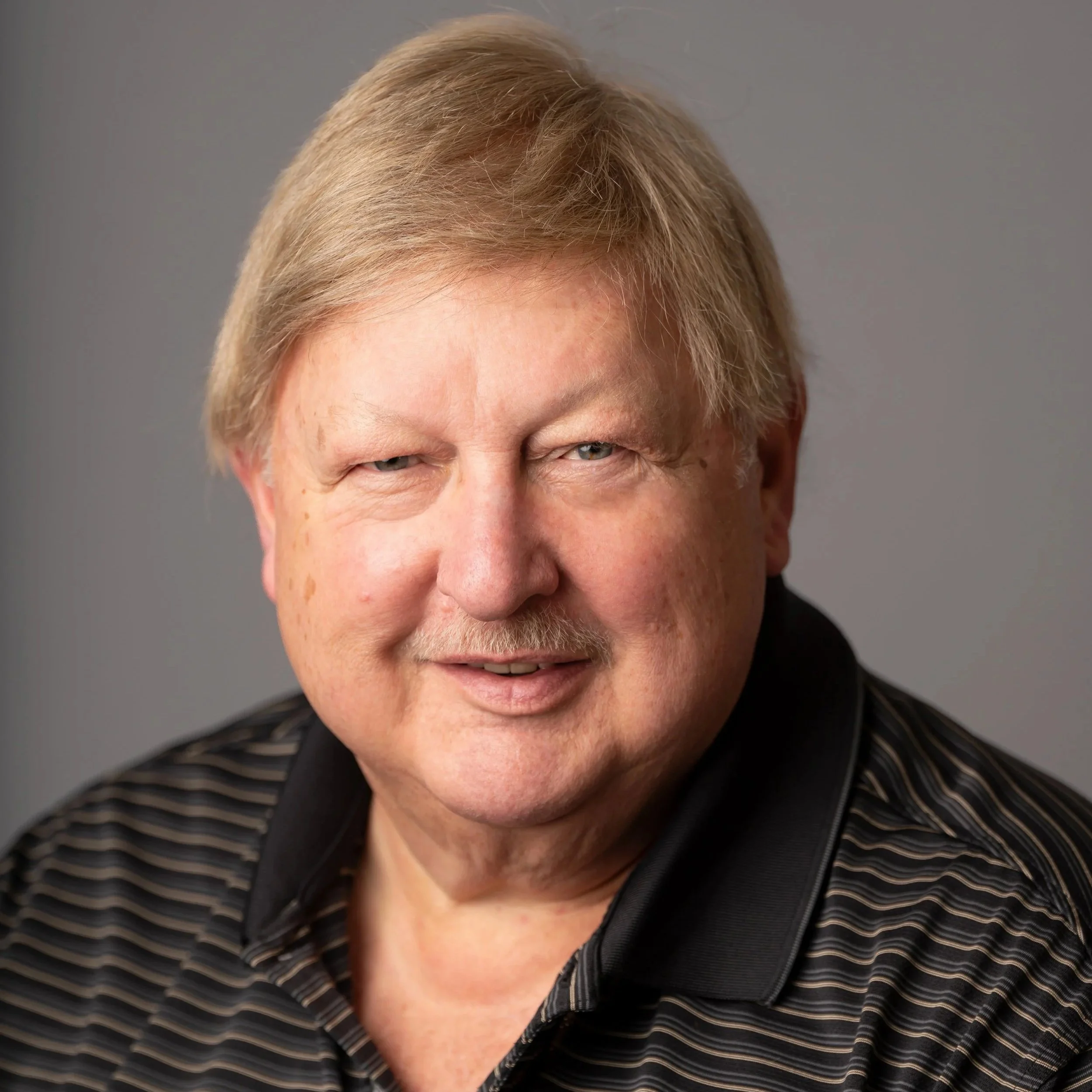Close-up portrait of a smiling older man with blonde hair, wearing a black striped polo shirt, against a neutral gray background.