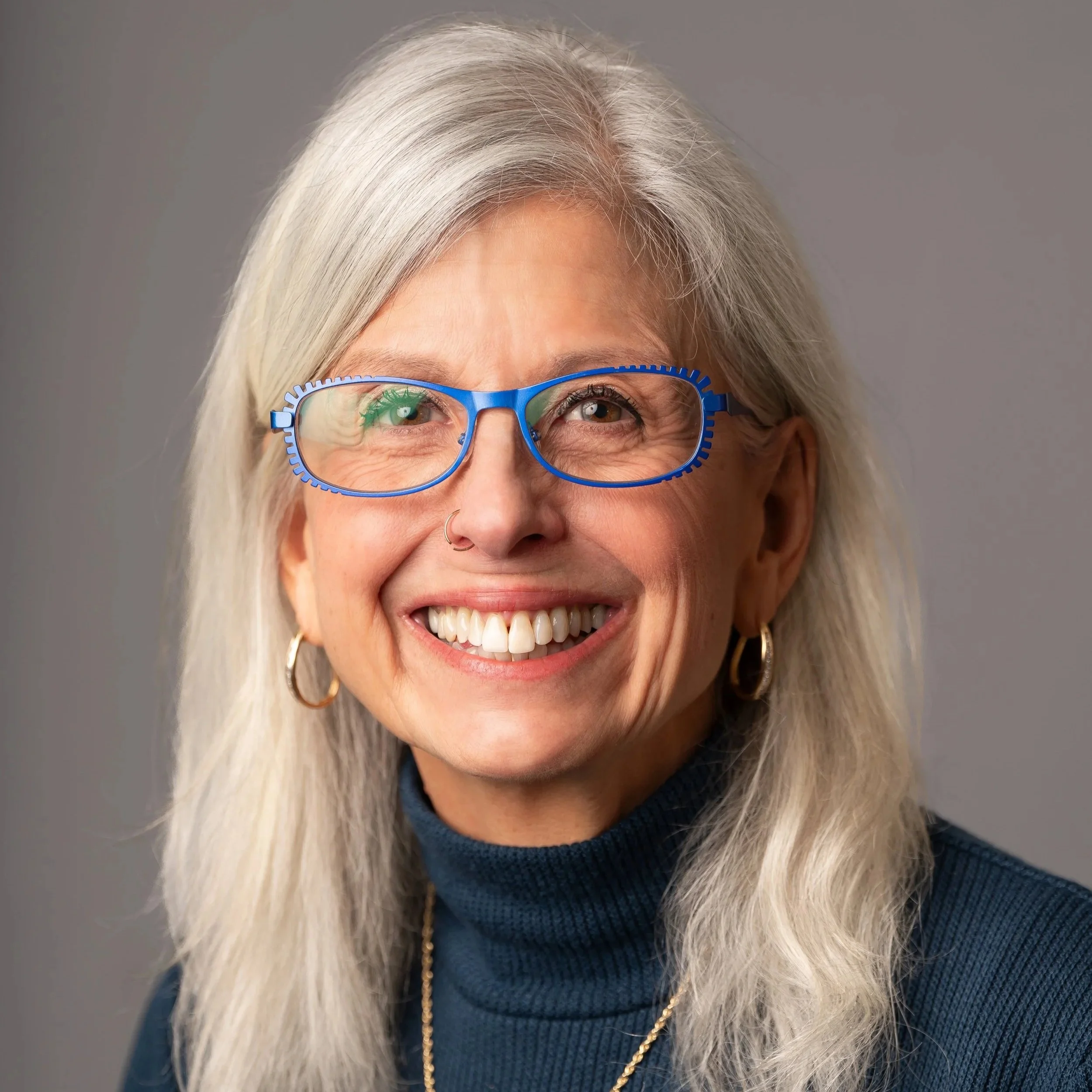 A smiling woman with long gray hair, wearing blue glasses, gold hoop earrings, a gold necklace with a cross, and a navy blue turtleneck, standing against a gray background.