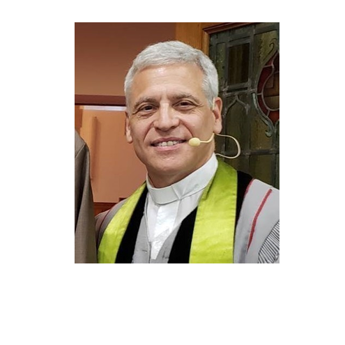 Man wearing a clerical collar and a bright green stole, smiling, with a headset microphone, standing indoors.