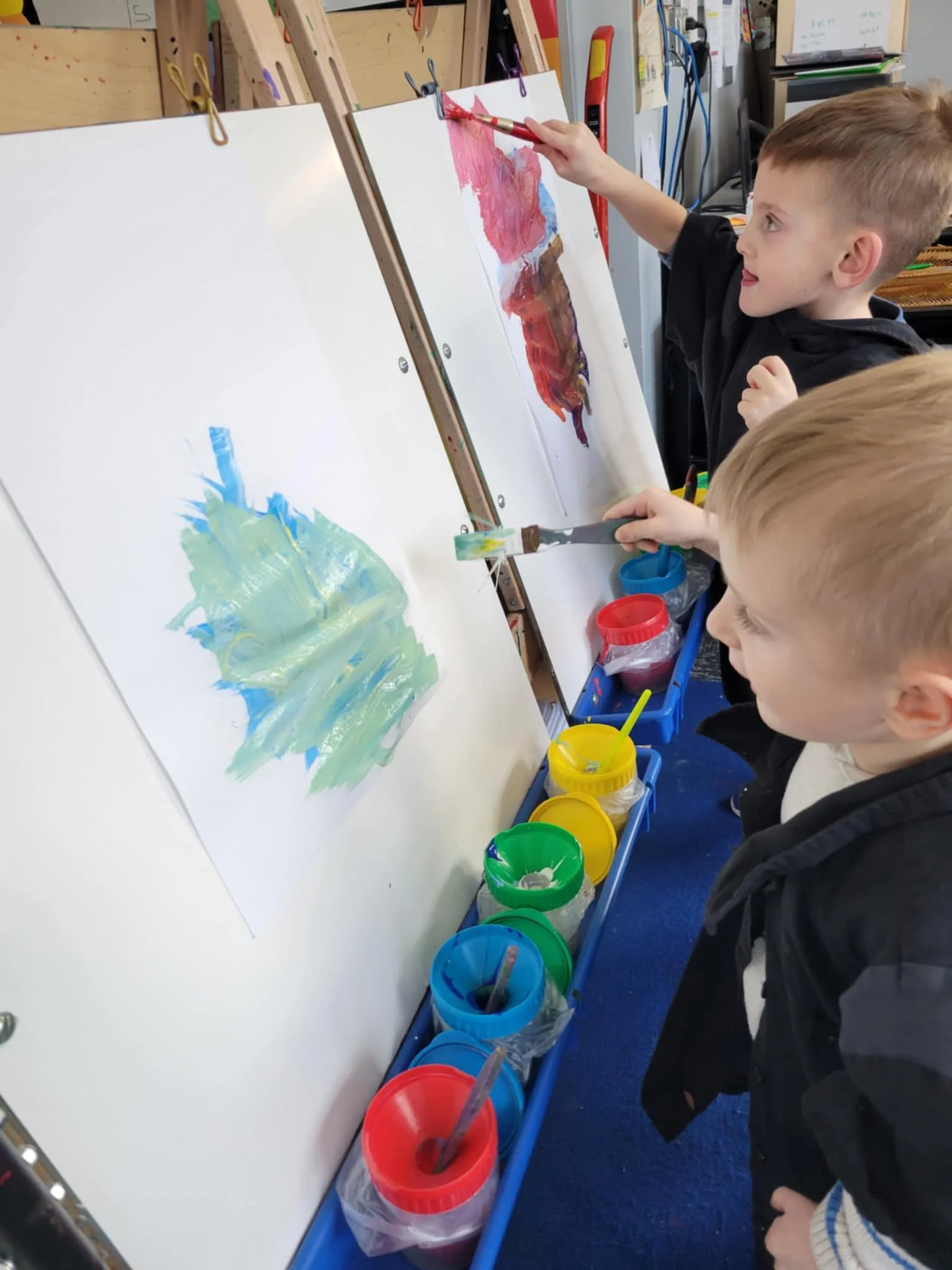 Side view of 2 students standing in front of an art easel.  Each student has several bright color paint options in their tray and are using paint brushes to paint a masterpiece.