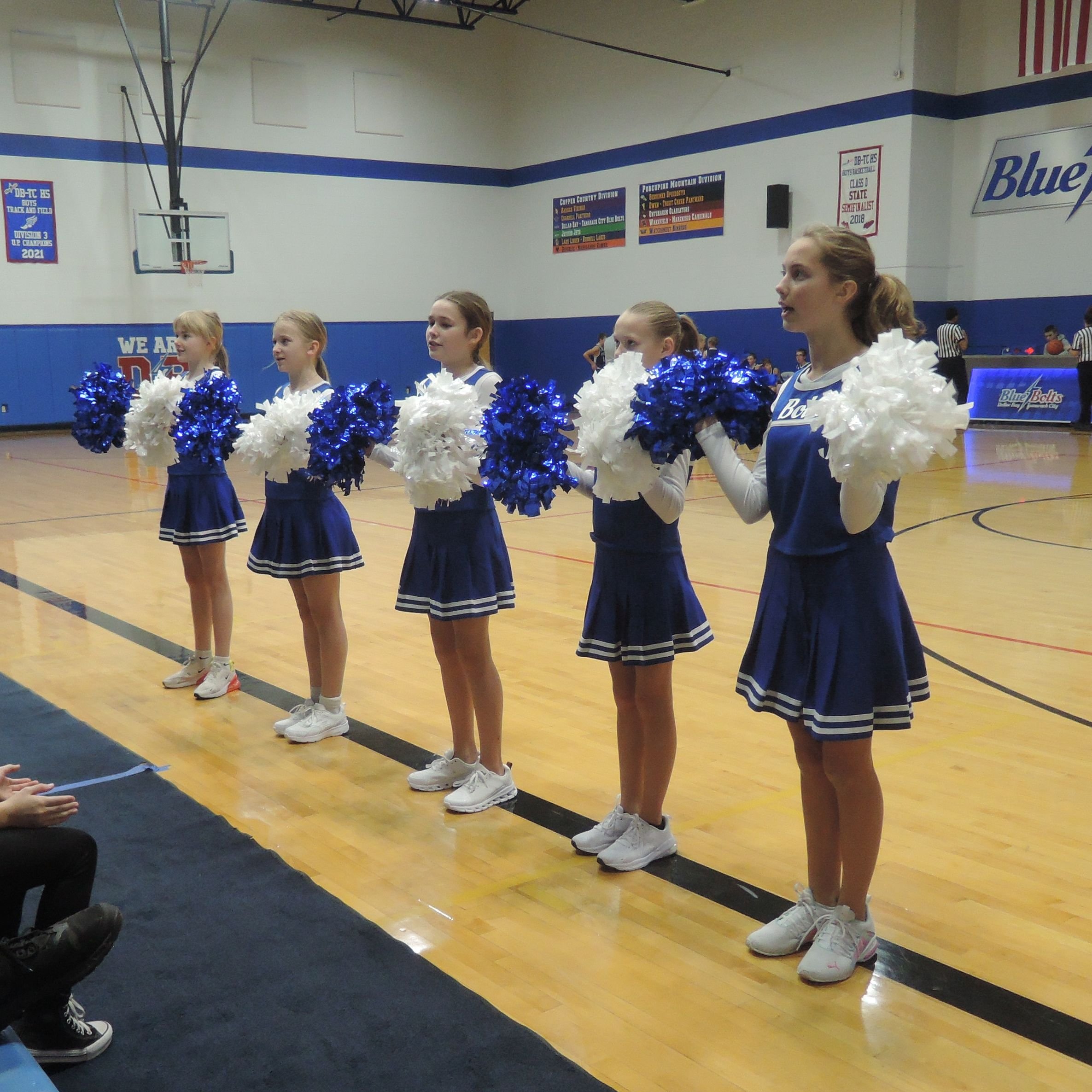 Five junior high cheerleaders, lined up on the side lines with blue and white pom poms, leading the fans in a cheer.