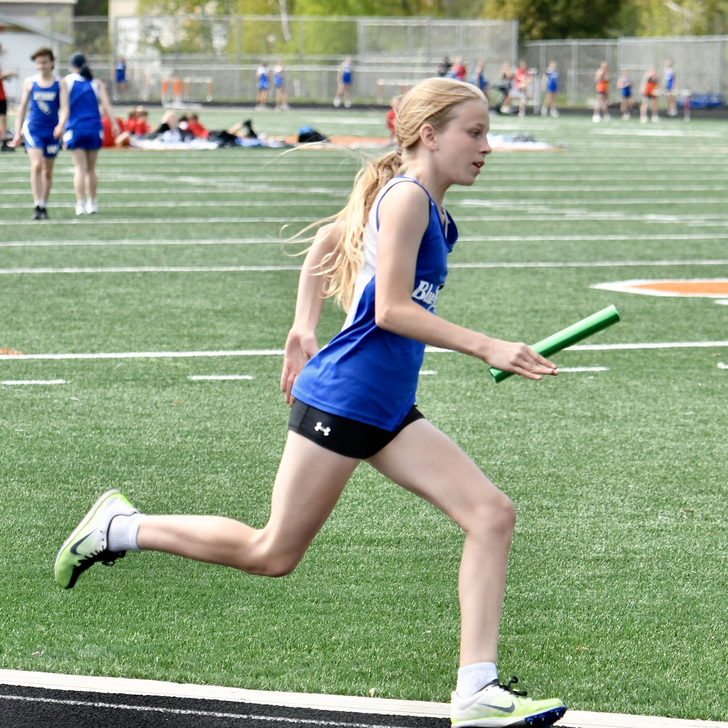 A junior high girl in full stride with a relay baton in her hands on the back curve of the track.