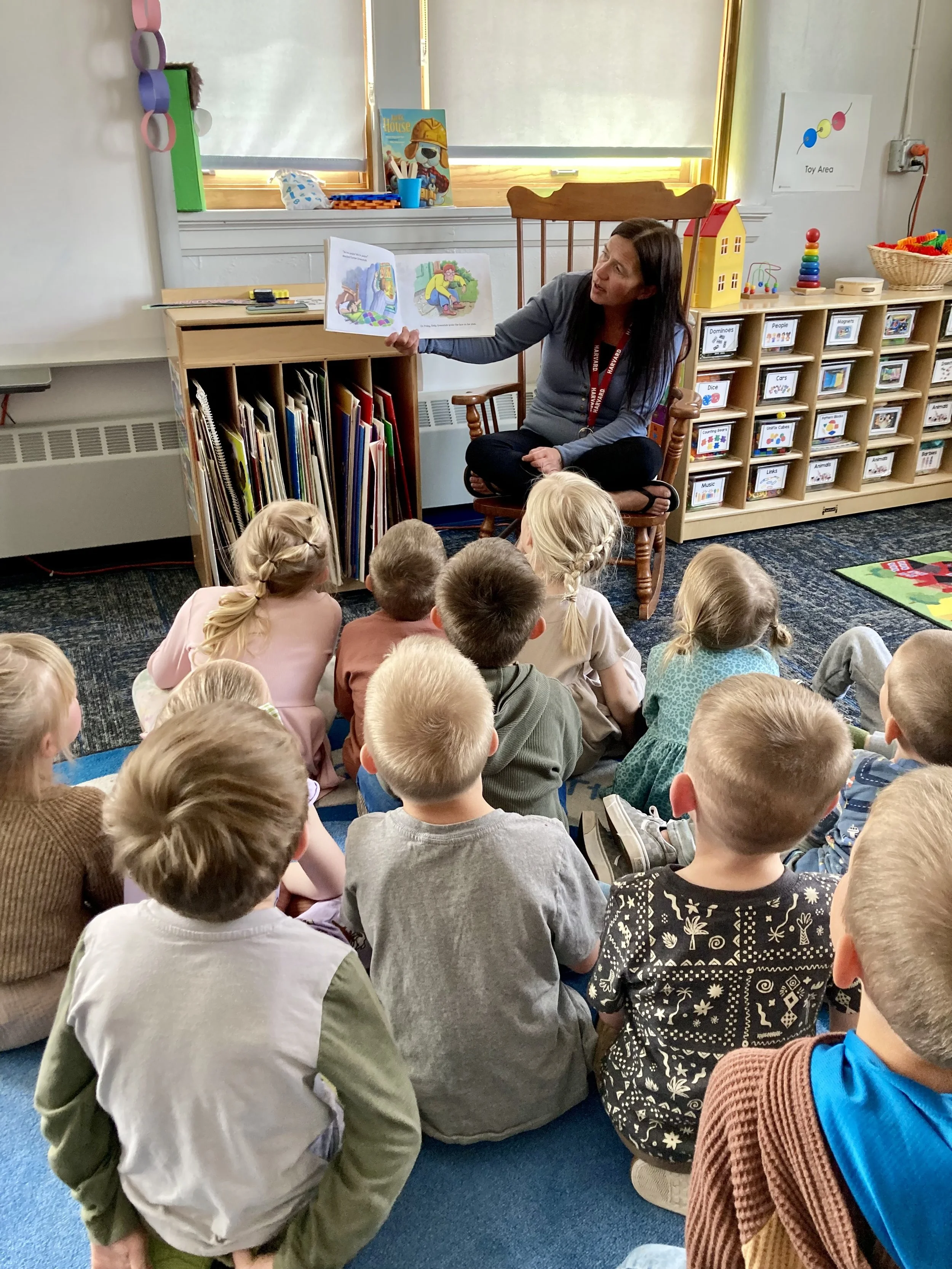 In the background is an assistant teacher sitting in a rocking chair facing the class and reading a book.  In the foreground is the back of the heads of a dozen preschool children attentively listening to the story.