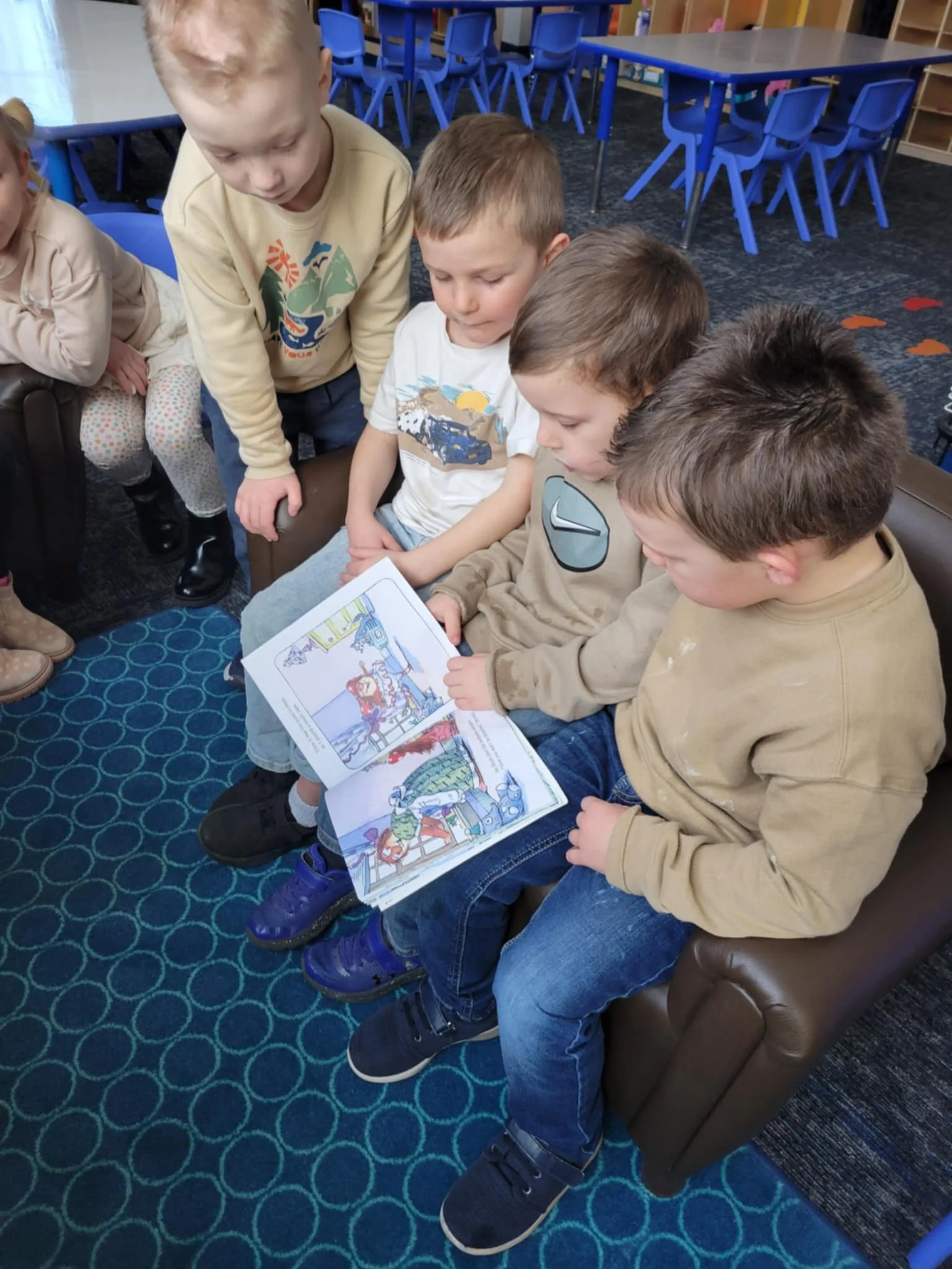 Three preschool students sharing a small brown leather  sofa.  The middle student has an open book in his lap.  All three are looking intently at the book toghether.