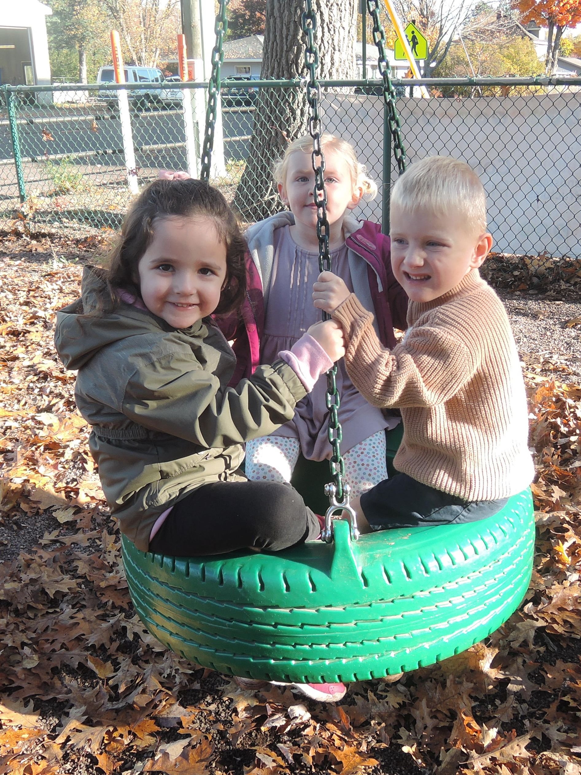 Three preschool students on a green tire swing in mid fall, smiling at the camera, with a think bed of fallen leaves below them.