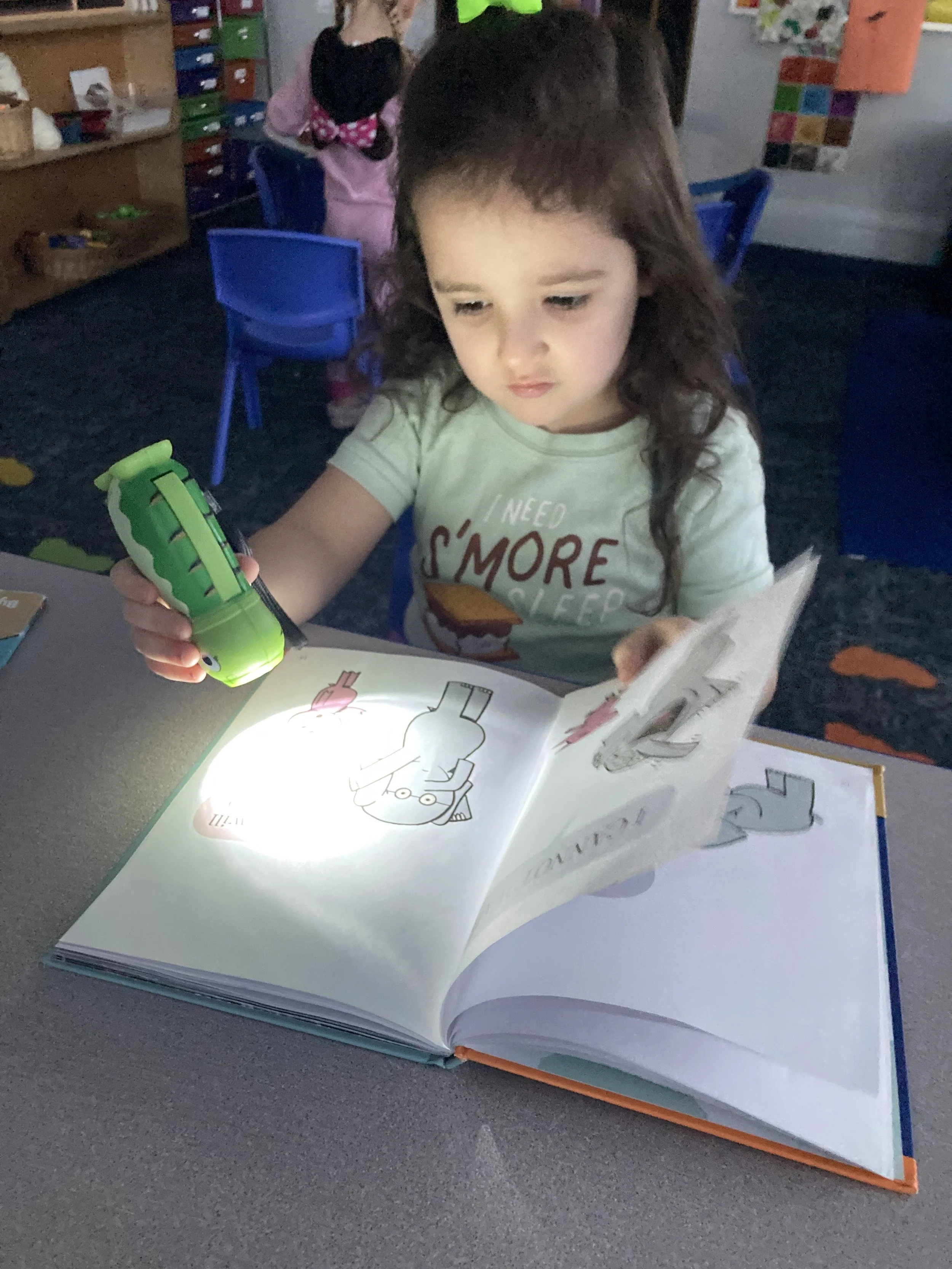A preschool student with a flashlight shining on a story book in a somewhat darkened room.