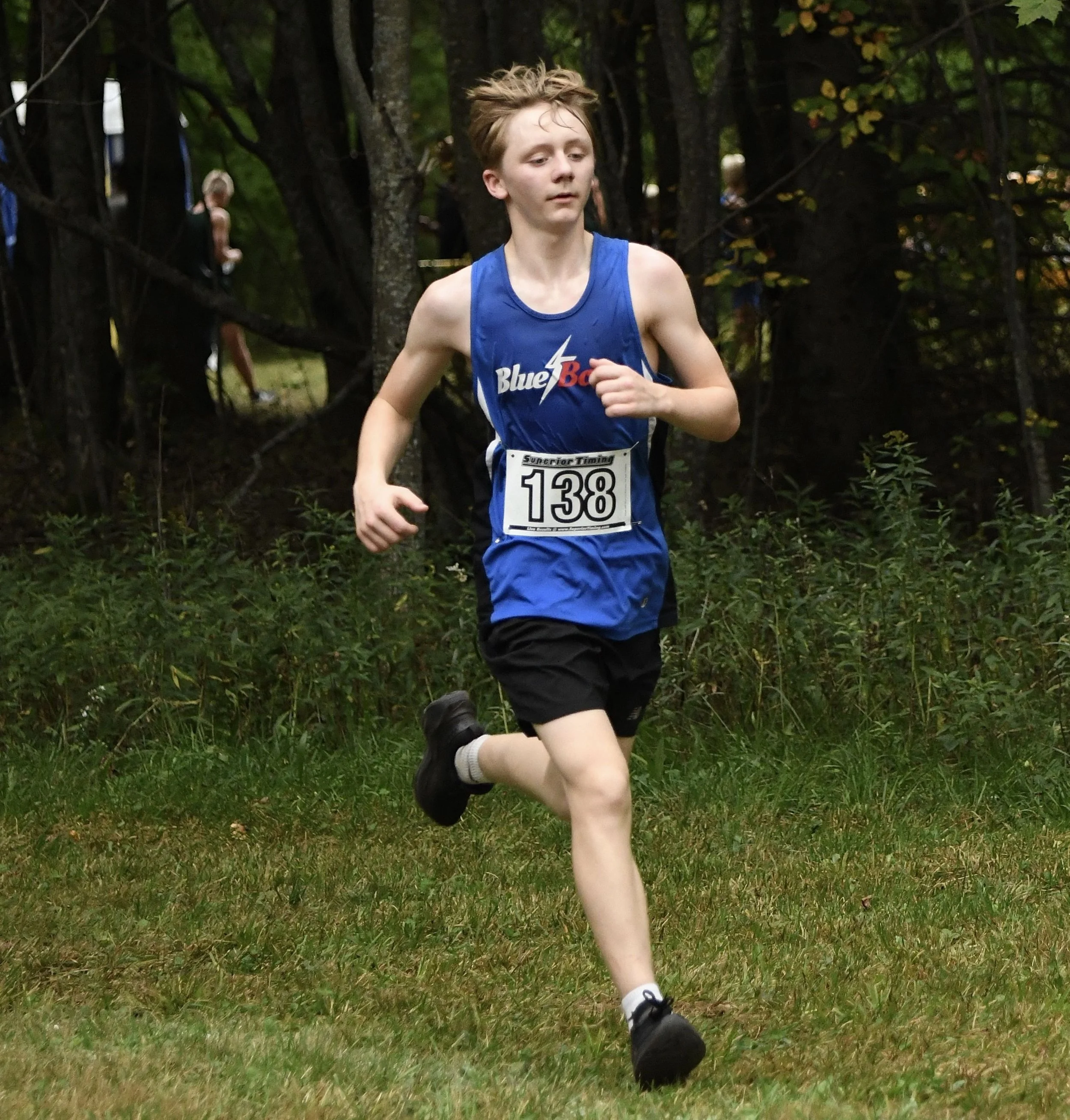 A varsity male cross country runner in full stride running on a green field with trees in the background.