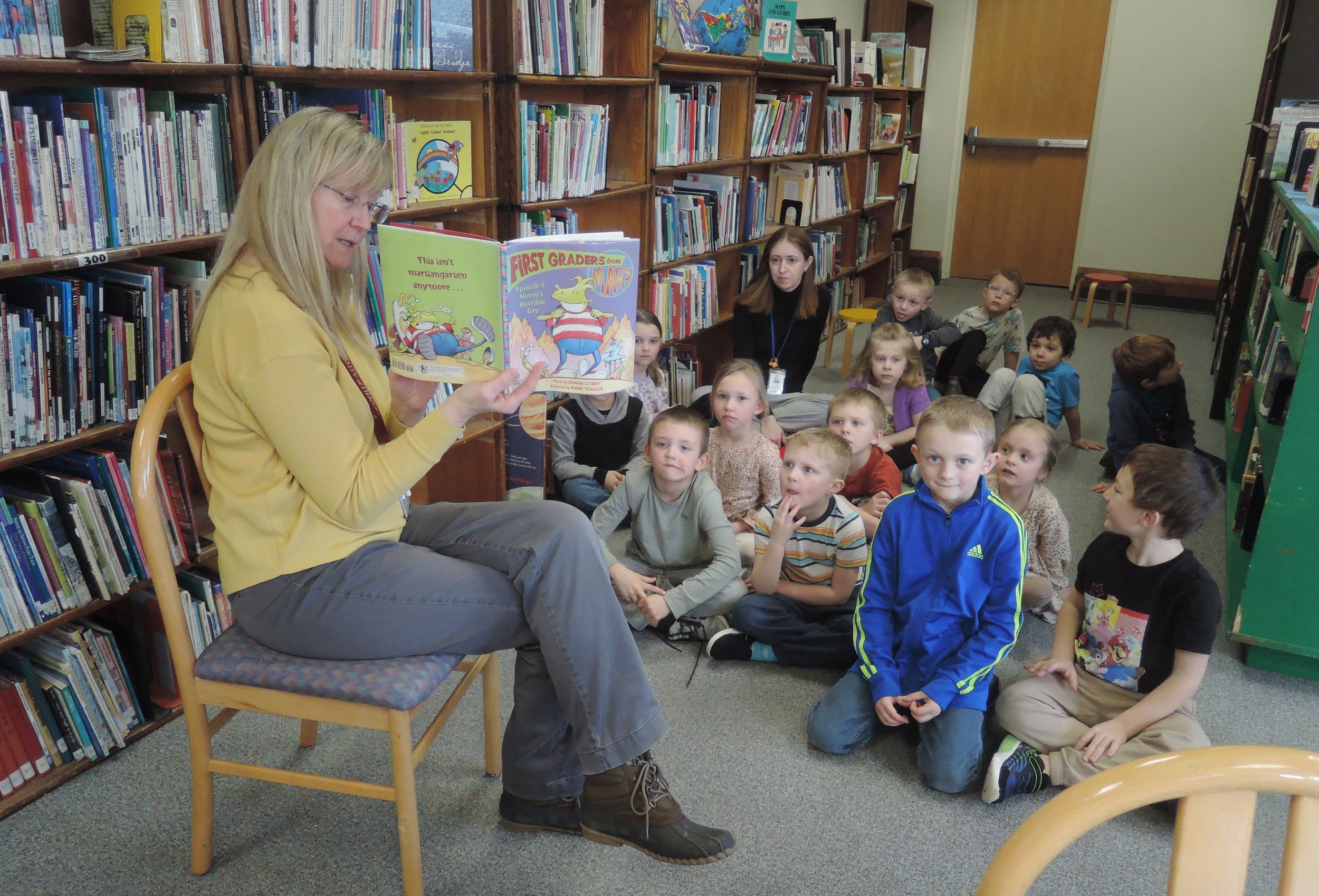 In the foreground is the librarian, sitting in a chair, legs crossed, displaying a book she is reading.  In the background is an elementary class, sitting cross legged on the floor,  attentively listening to the story.