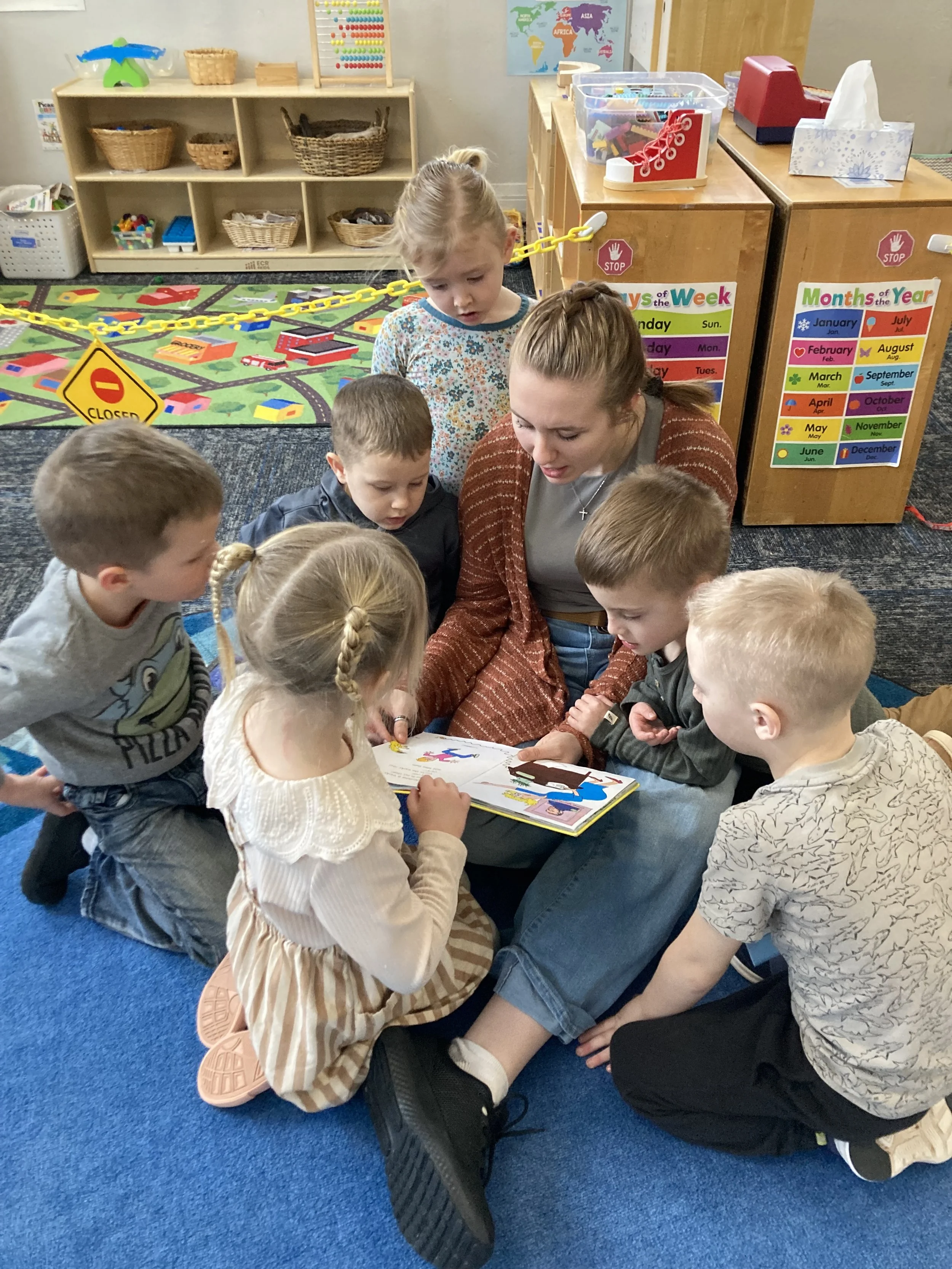 A preschool assistant sitting on a blue carpeted floor, reading a book to a half dozen students sitting all around her.