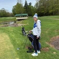 A female member of the golf team posing in front of her bag of golf clubs.  In the background is a pond, and a bridge spanning the stream that feeds the pond.