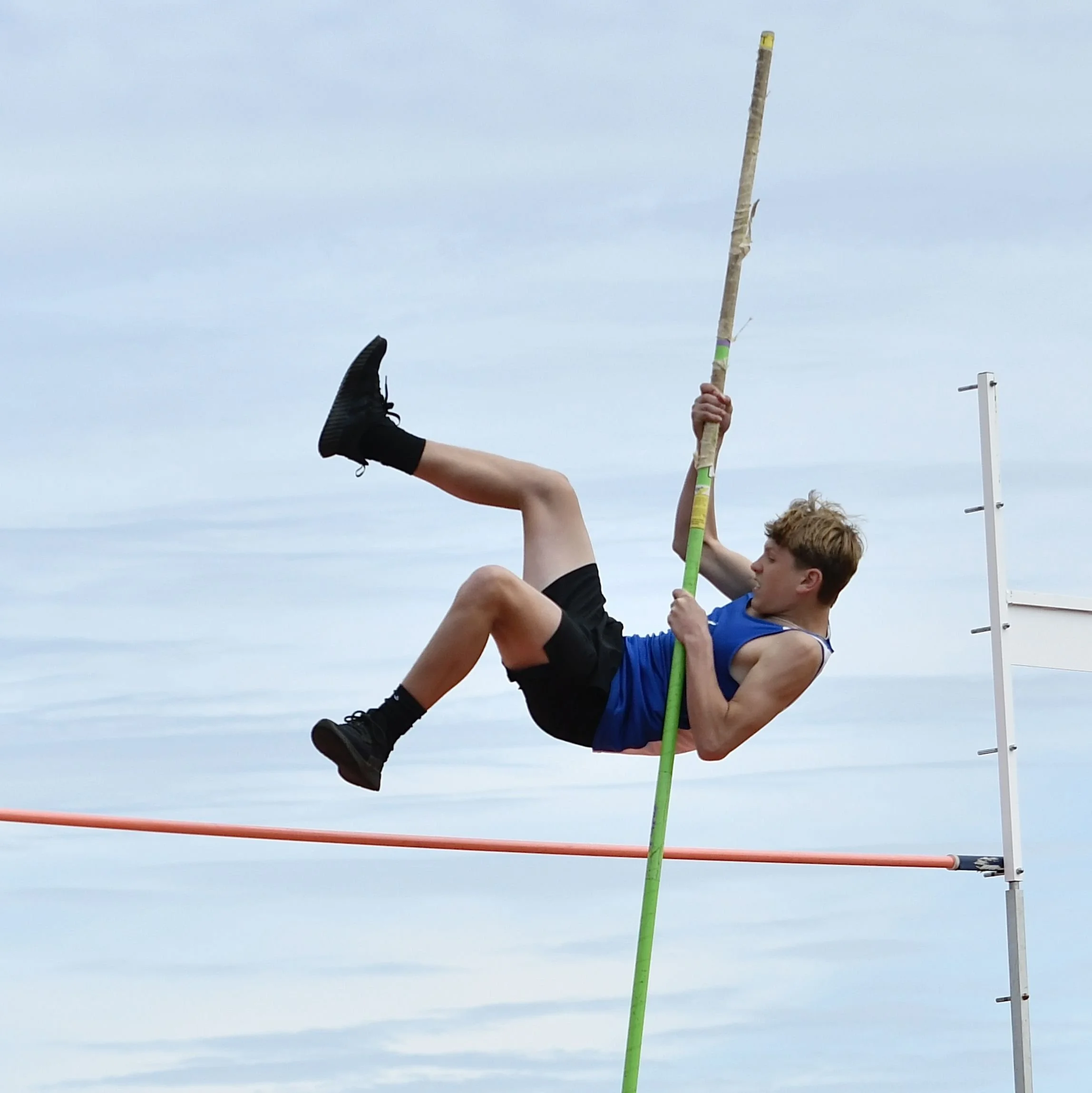 A junior high boy pushing on his pole to lift himself over the crossbar  in completion of a very successful pole vault.