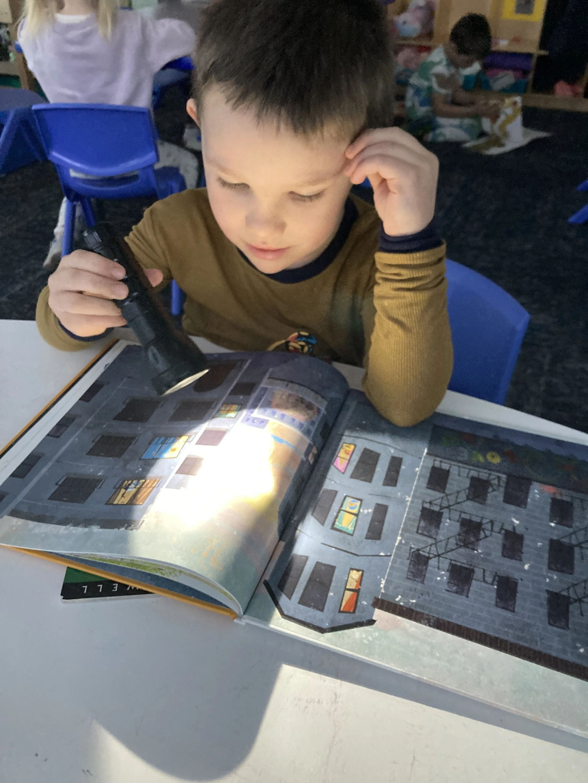 A preschool student with a flashlight shining on a story book in a somewhat darkened room.