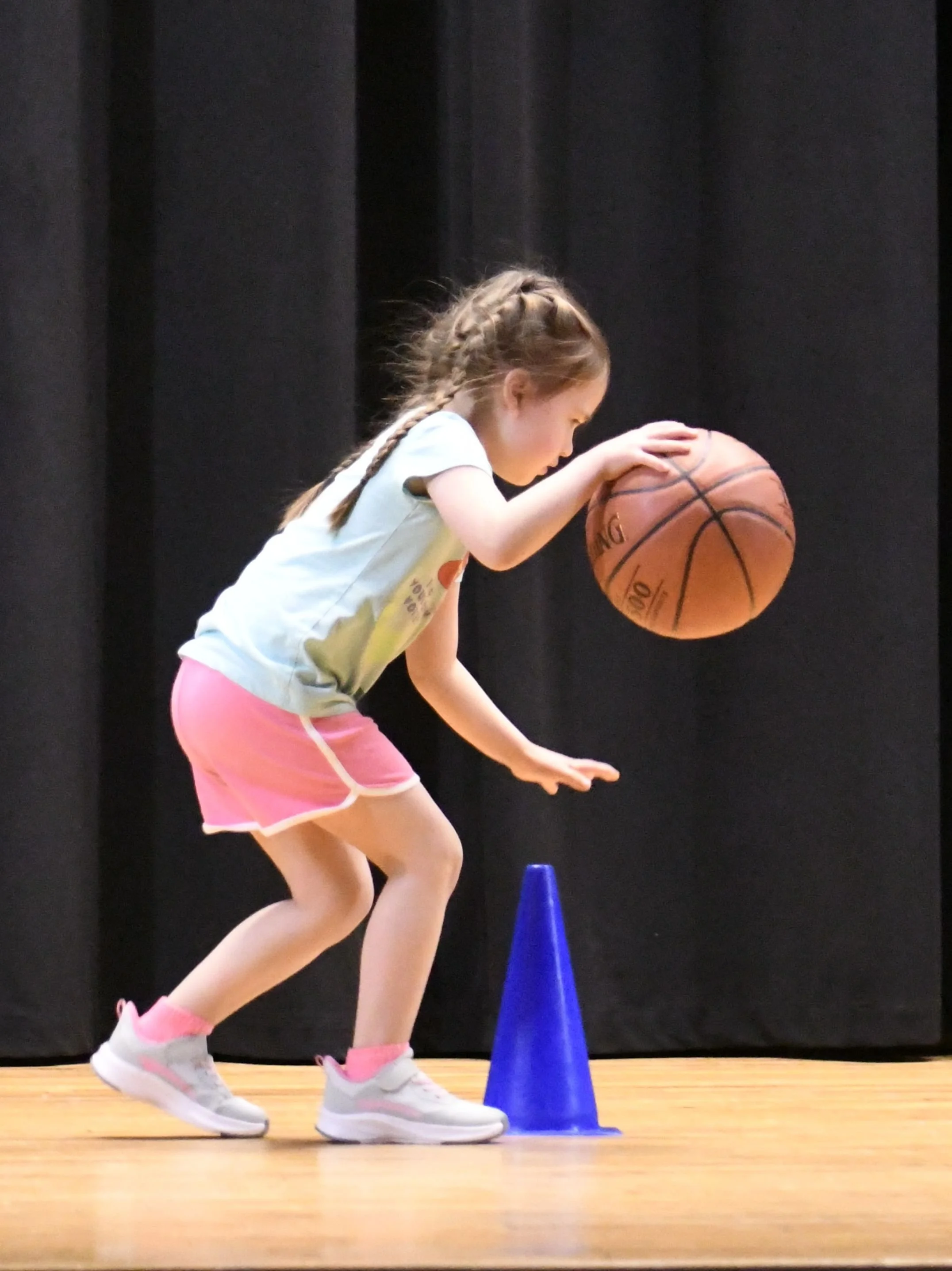 A kindergarten student on stage at a talent show, showing her skills in dribbling  basketball around a cone.