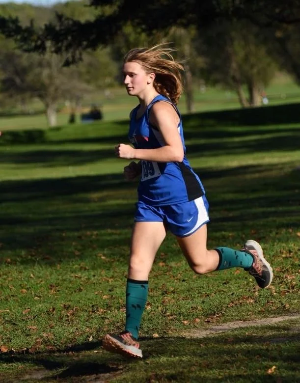 A Lone high school female running at a brisk stride alongside a well maintained grass field. with scattered trees,