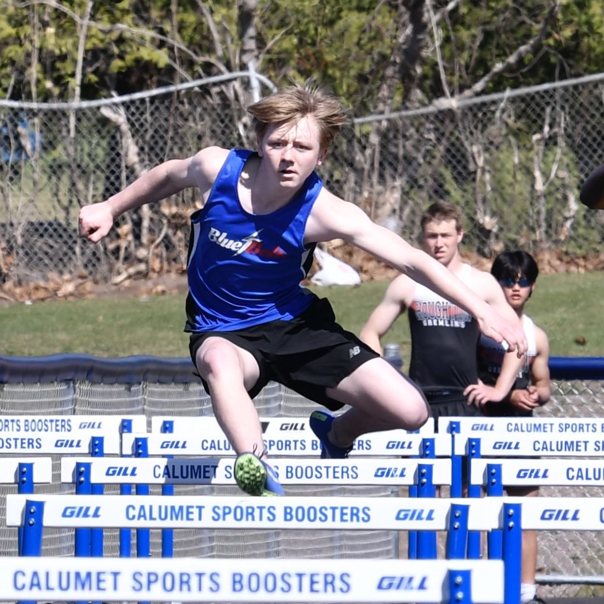 A high school male facing the camera, in mid stride over a hurdle, one leg forward and the trail leg bent.