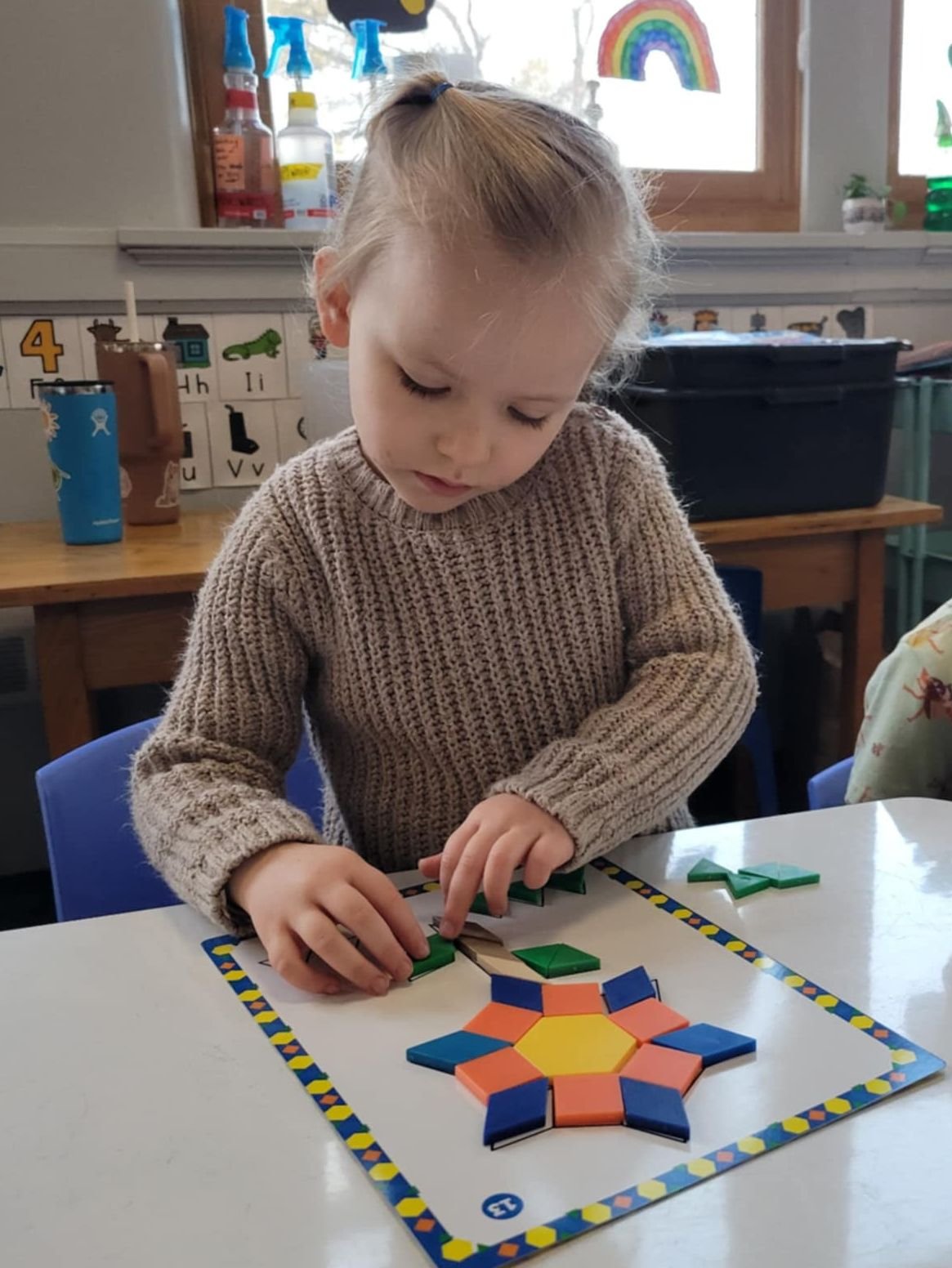 A preschool student standing over a table, putting together a picture of a beautiful flower suing colorful manipulative shapes.